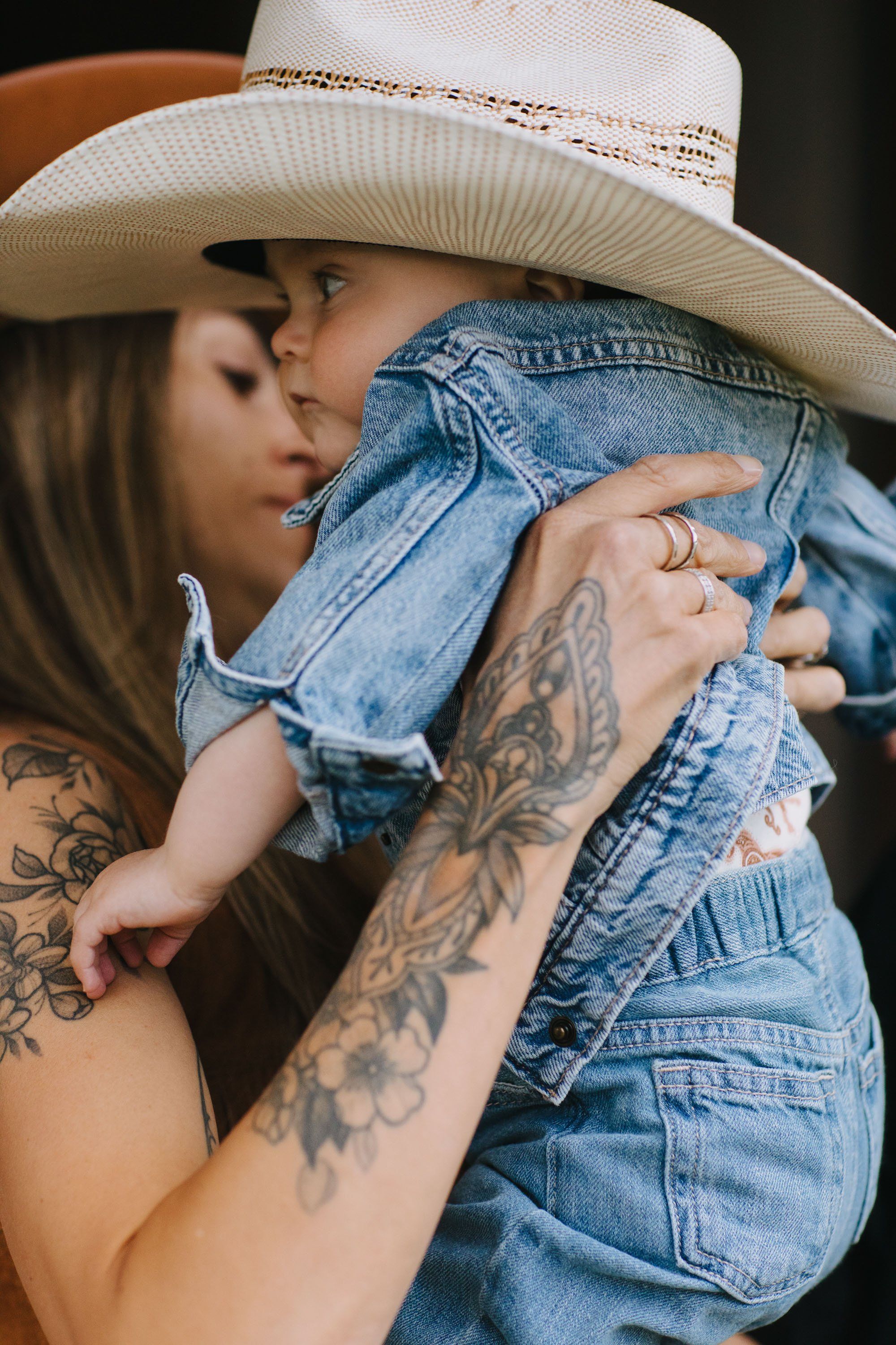 Woman with visible tattoos embraces child wearing a cream cowboy hat against a dark background