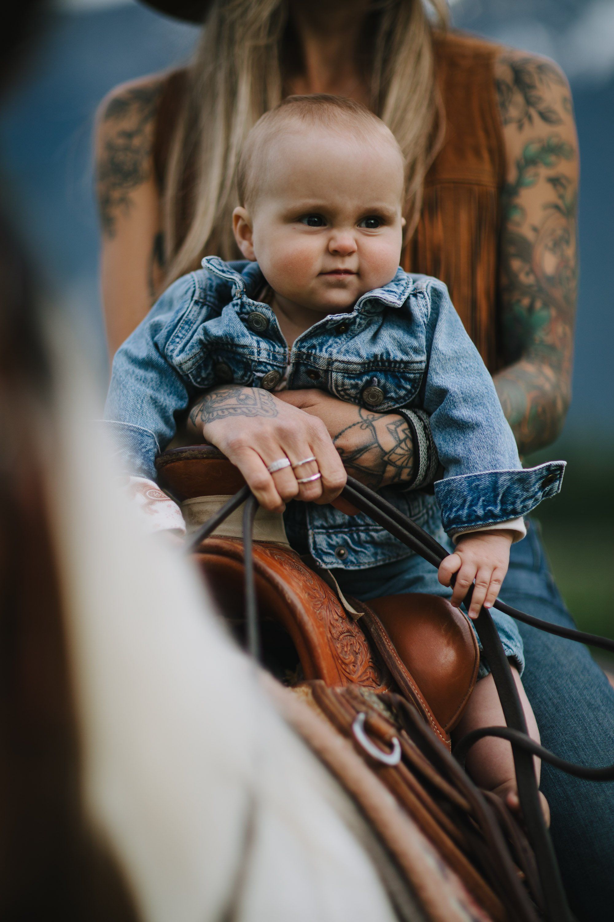 Tattooed parent holding a smiling infant in denim on horseback, warm natural light, mountains blurred behind
