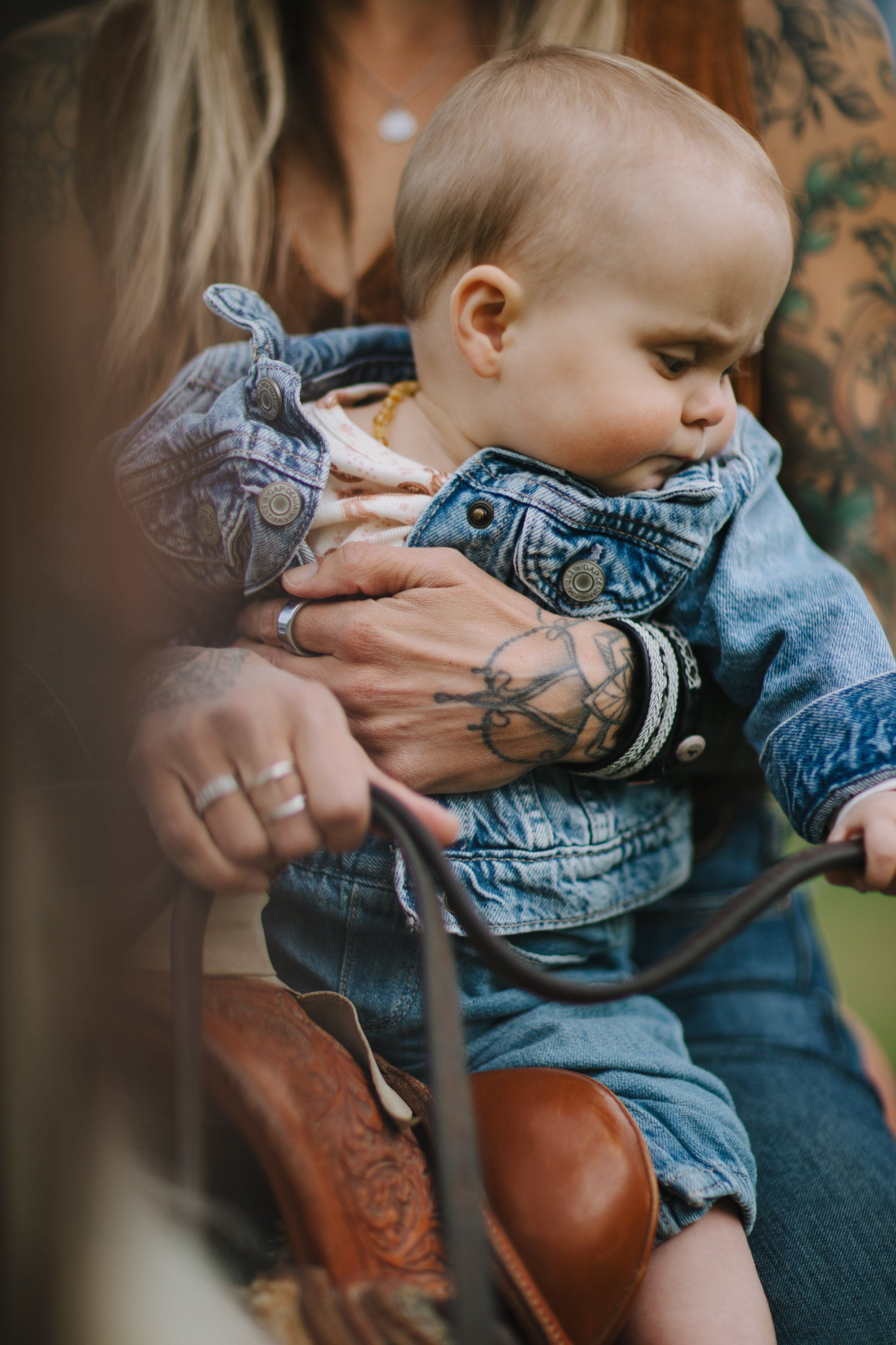 Tattooed parent holding toddler in denim jacket, soft natural light filtering through trees in outdoor setting