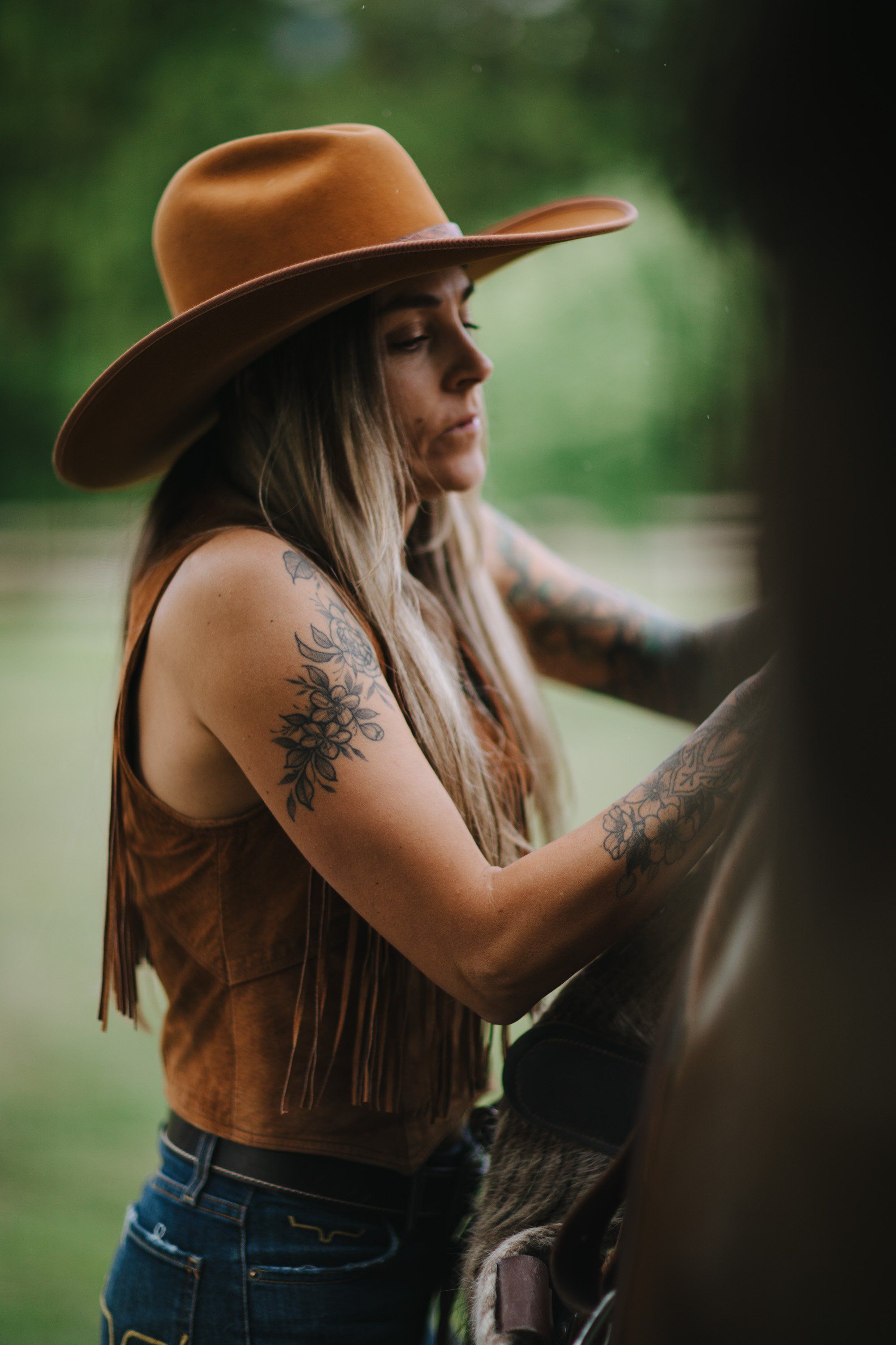 Woman in tan cowboy hat and brown suede vest with visible arm tattoos, posed against wooden post in forest setting