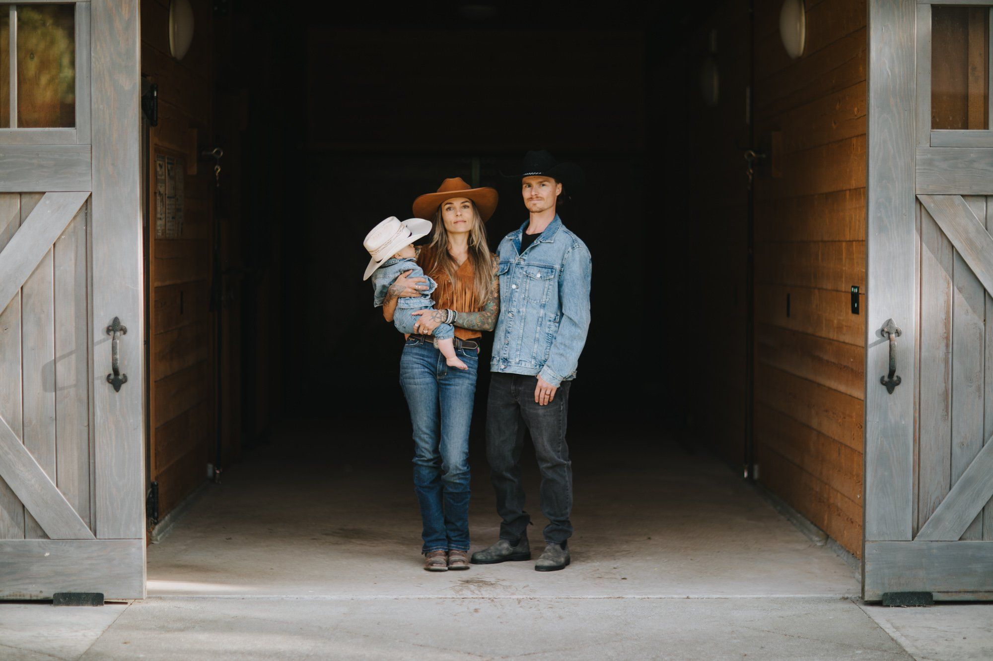 Family of three in cowboy hats standing in a sunlit barn doorway with warm golden light streaming across the wooden floor