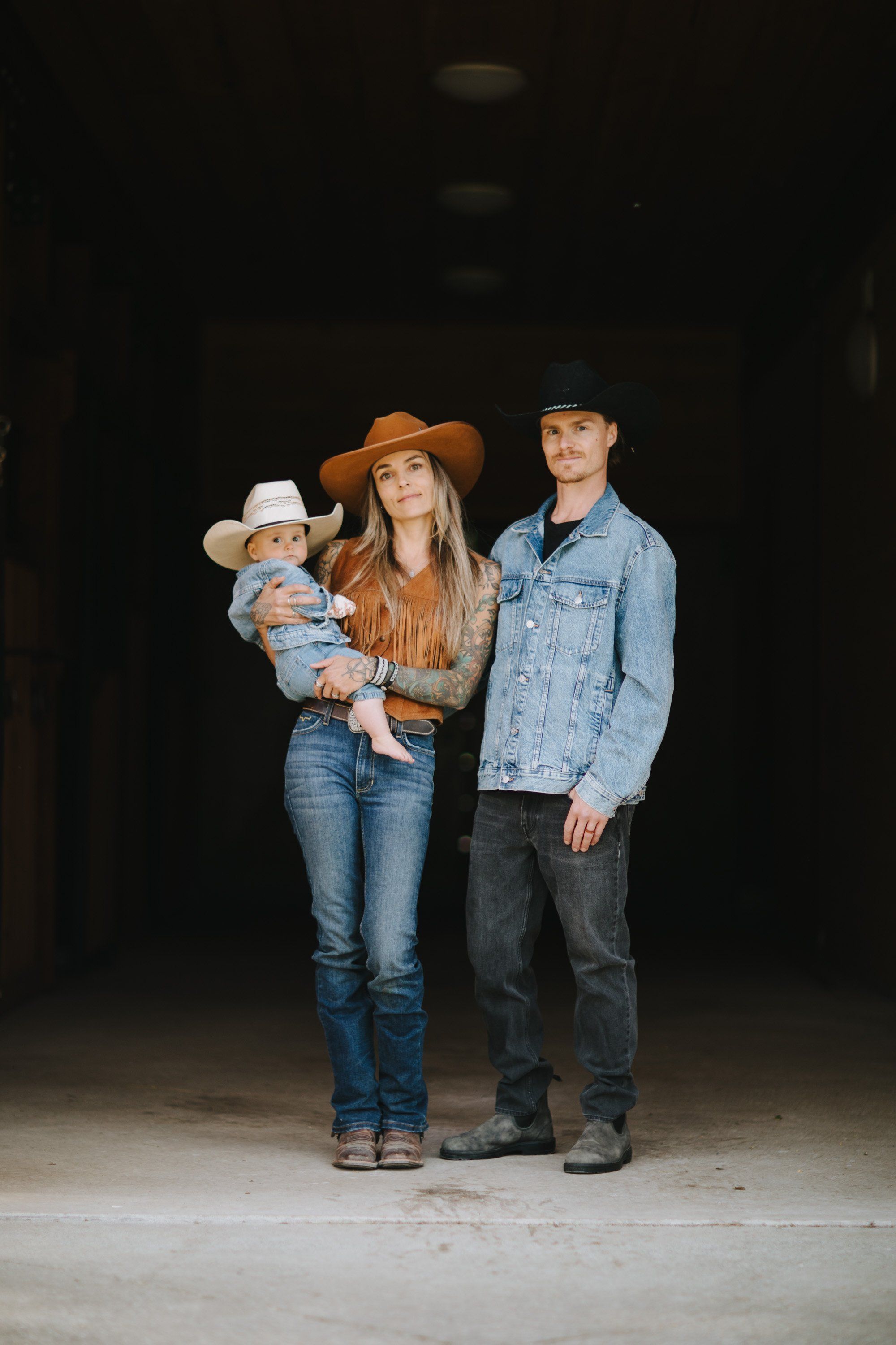 Family of three in cowboy hats posed against dark barn interior, warm studio lighting on concrete floor