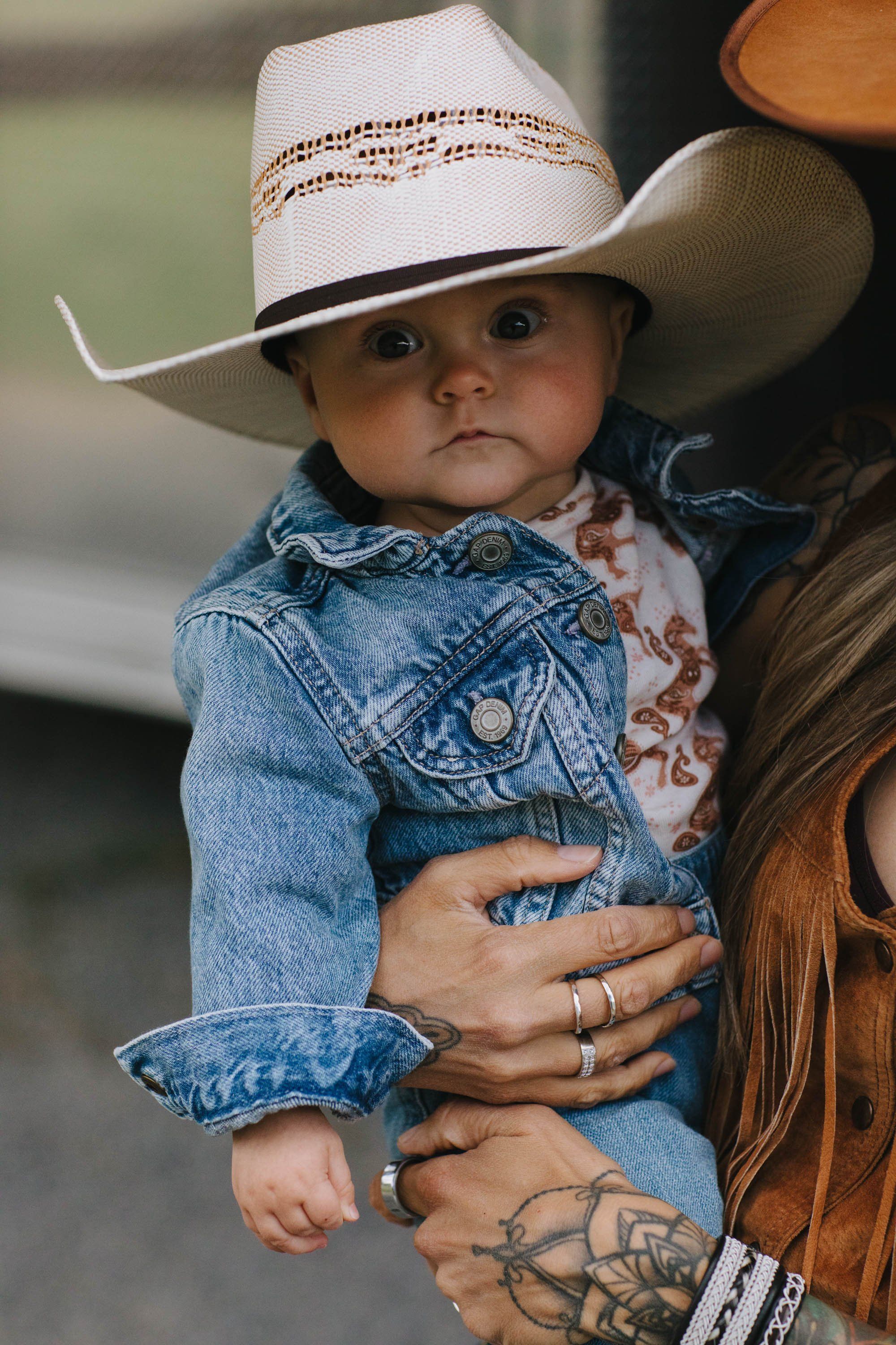 Toddler in denim jacket and white cowboy hat held by parent with visible tattoos and rings, soft natural light