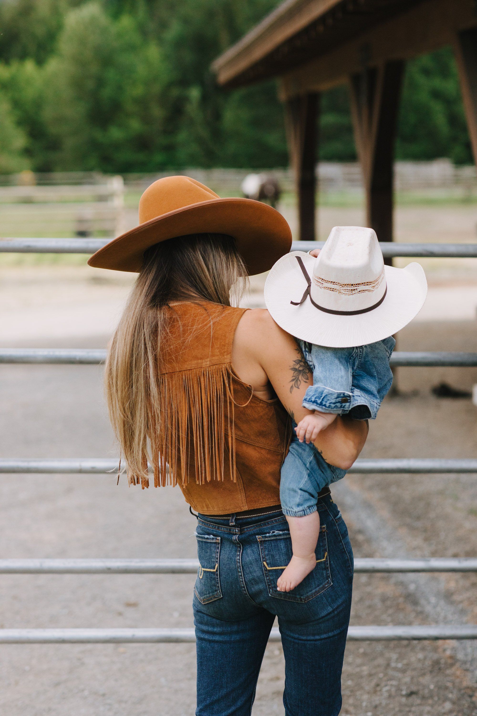 Woman in tan fringe jacket and brown cowboy hat holds toddler wearing white hat at rustic ranch fence