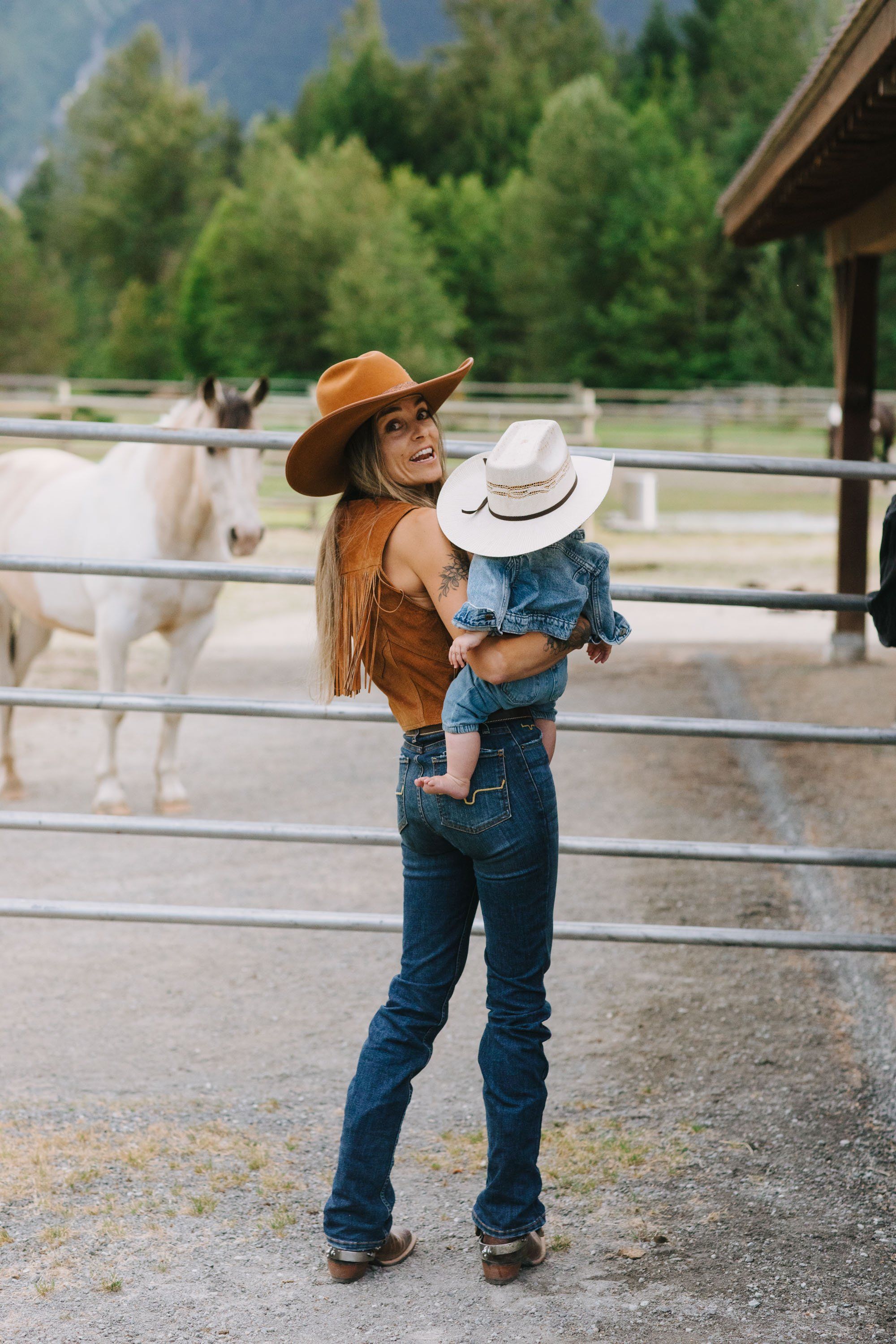 Woman and child in western wear stand at a ranch fence with horses and forested mountains behind them