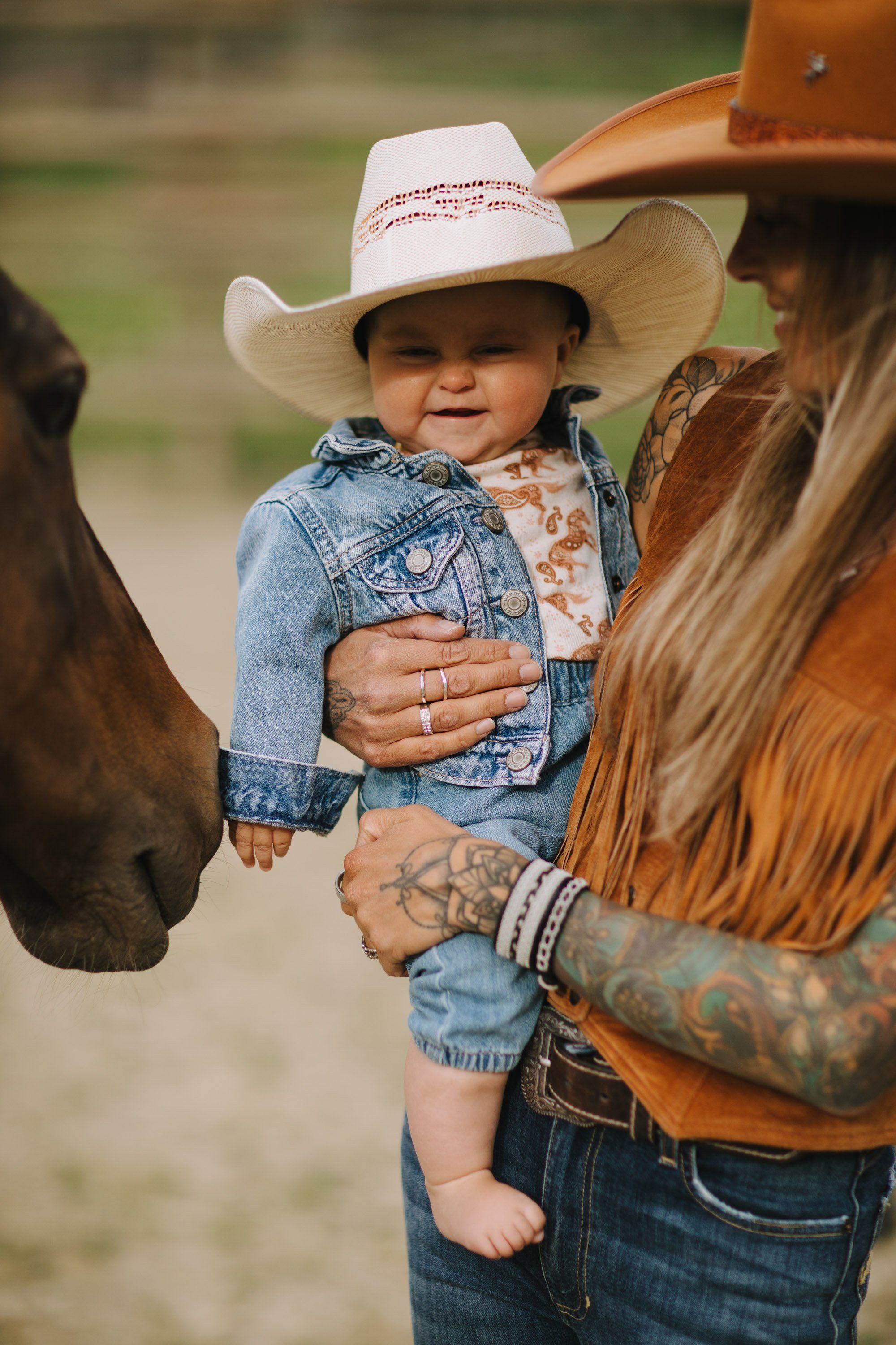 Toddler in cowboy hat and denim jacket held by tattooed adult near horse in golden afternoon light