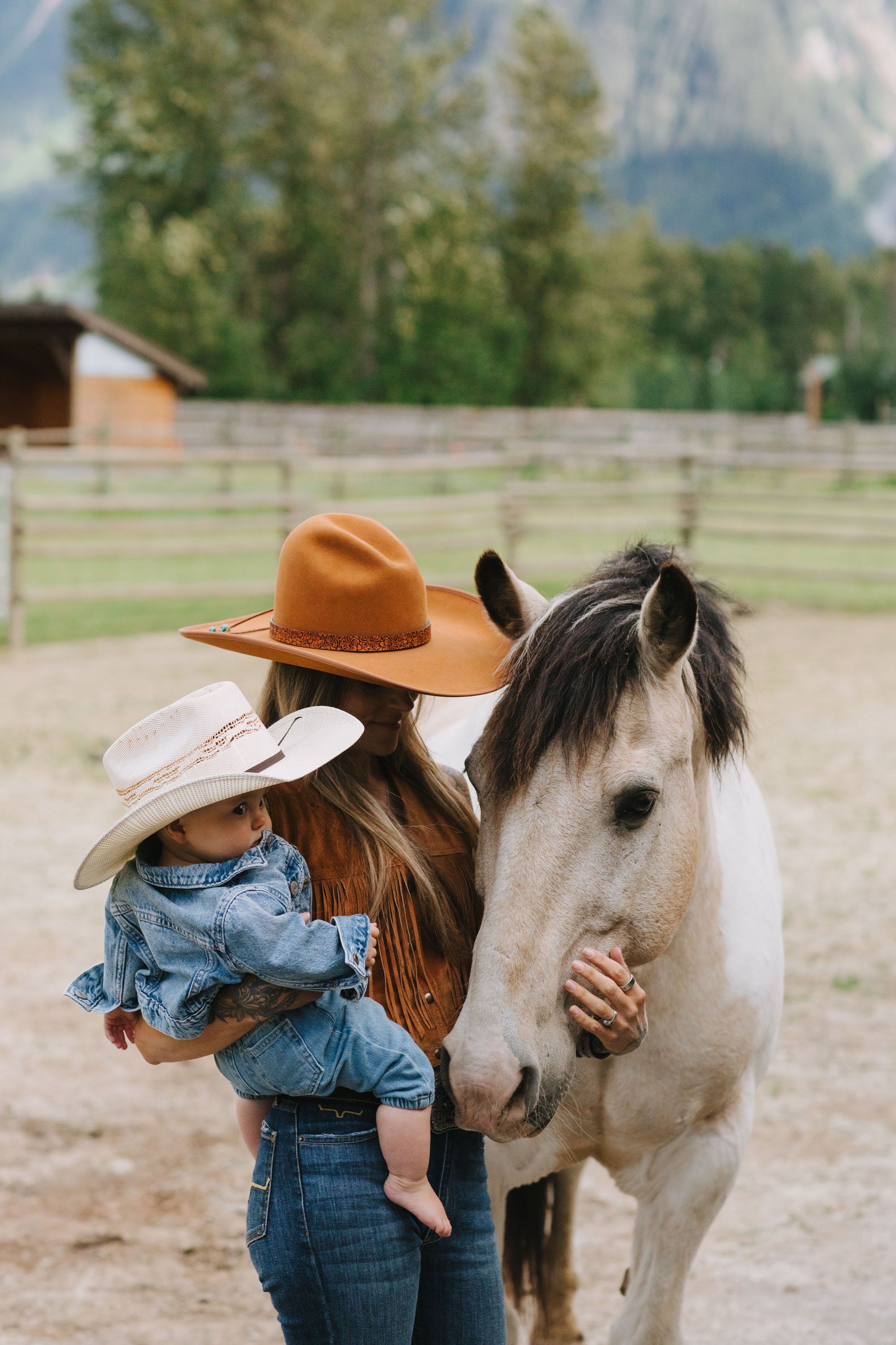 Girl in cowboy hat and denim embraces white horse in sandy arena with mountains and pine trees beyond