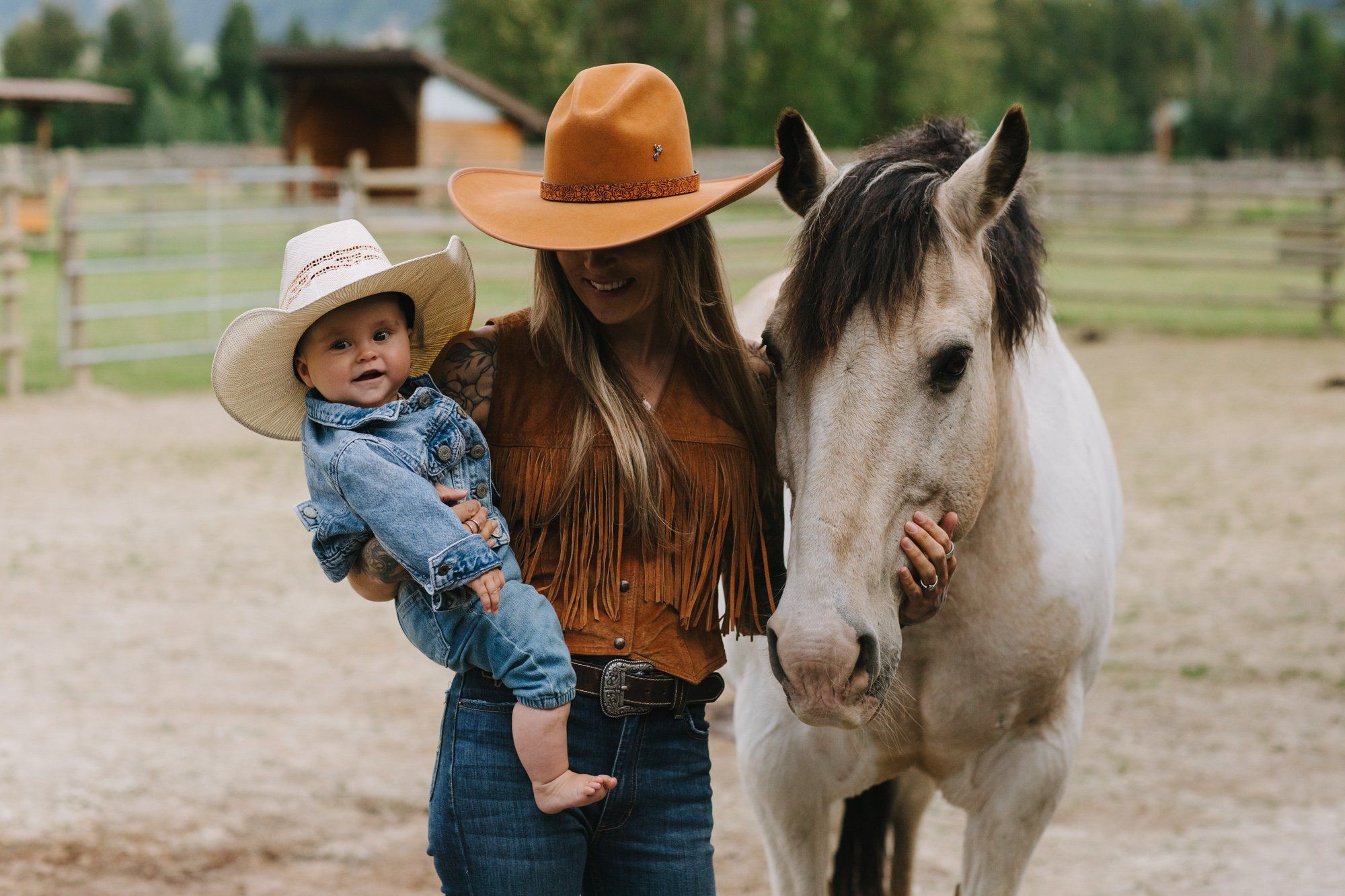 Woman in brown cowboy hat holding toddler in white hat beside white horse in rural ranch setting