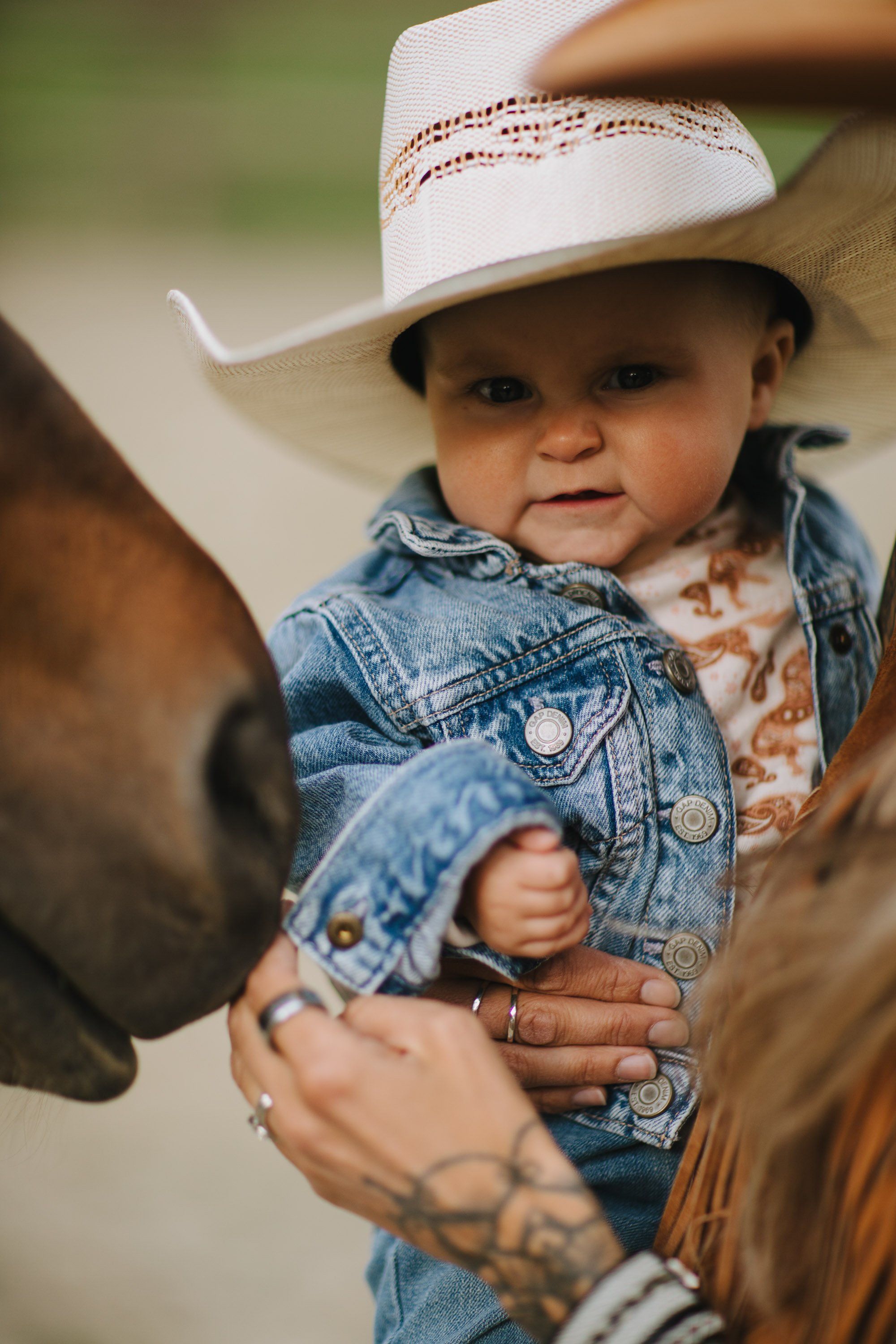 Toddler in white cowboy hat and denim jacket held by parent, soft natural light, warm countryside setting