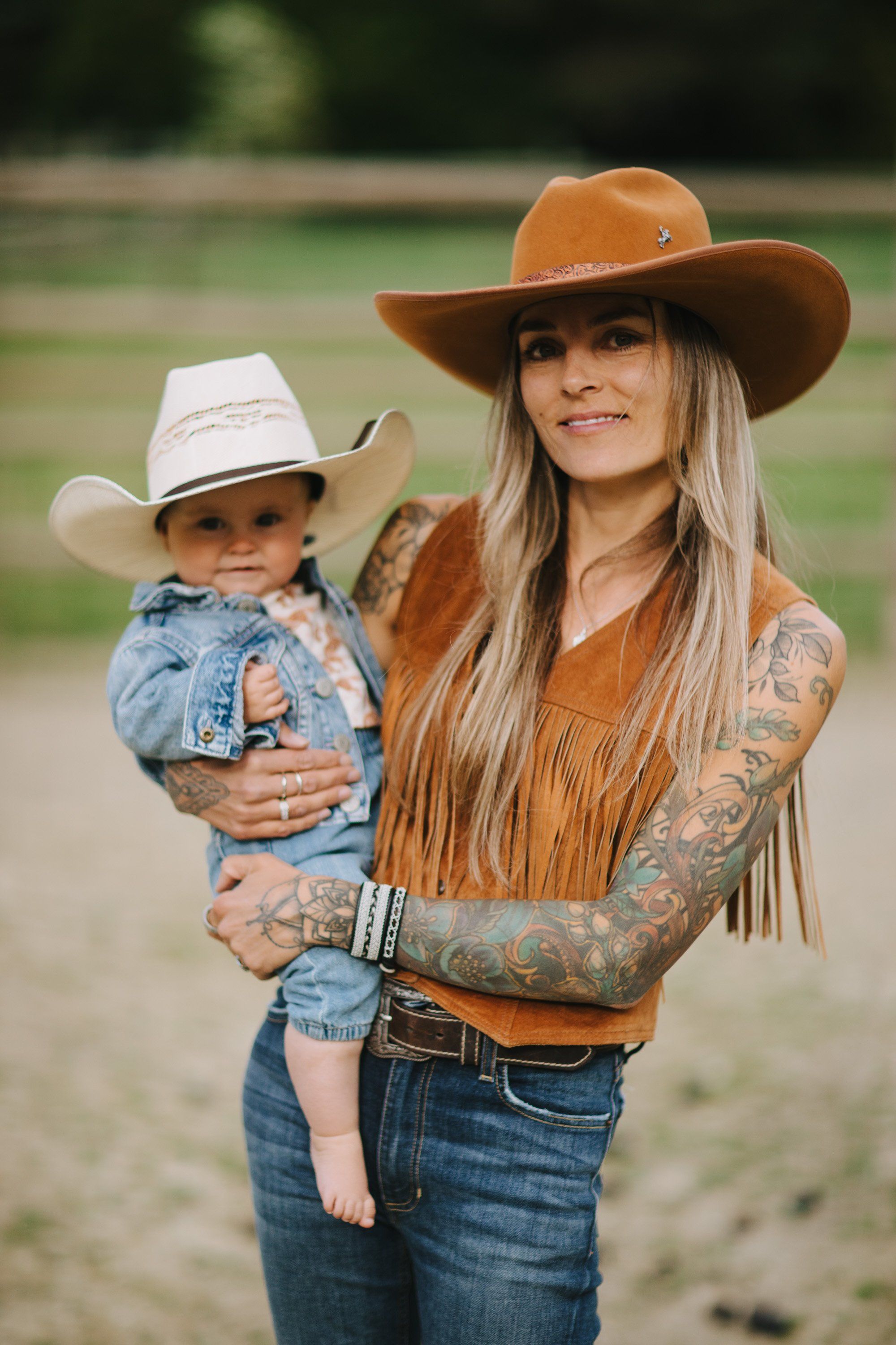 Woman in brown cowboy hat and fringe top holding toddler in white hat, both smiling at camera in rural setting