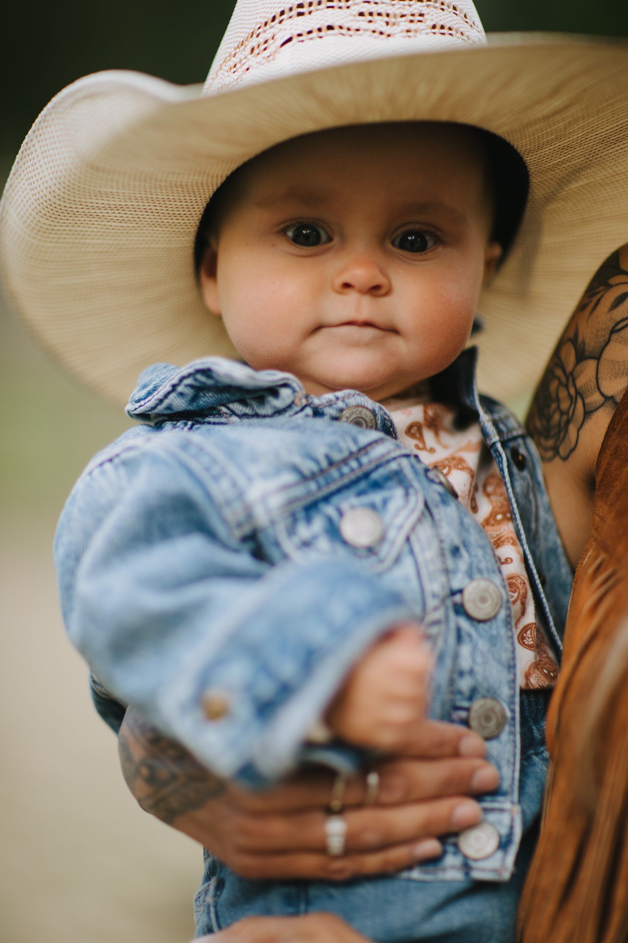 Toddler in denim jacket and cream cowboy hat held by parent with tattooed arm against soft green background