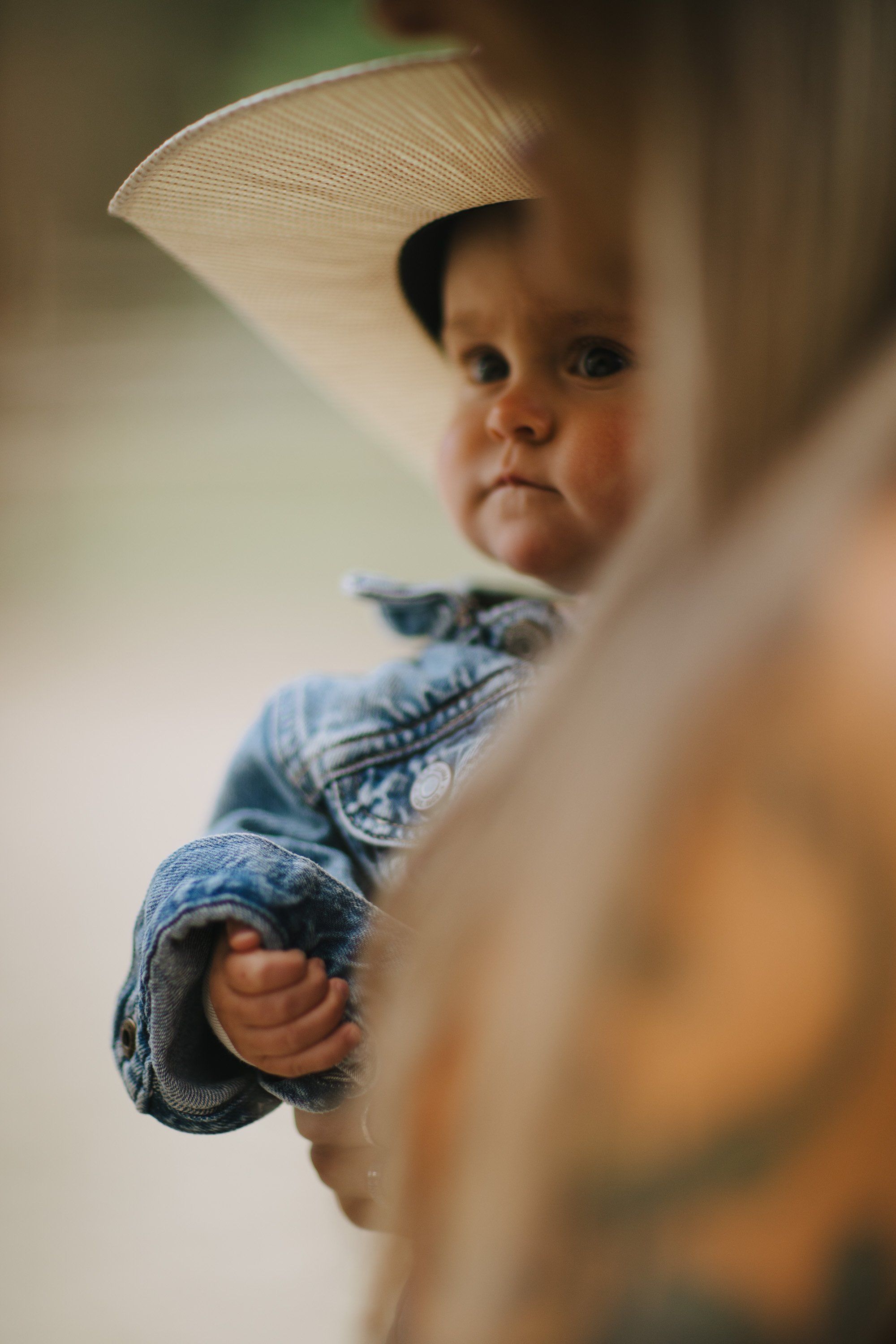 Toddler in denim peeking around a wooden post with curious expression, soft natural light and warm blur in foreground