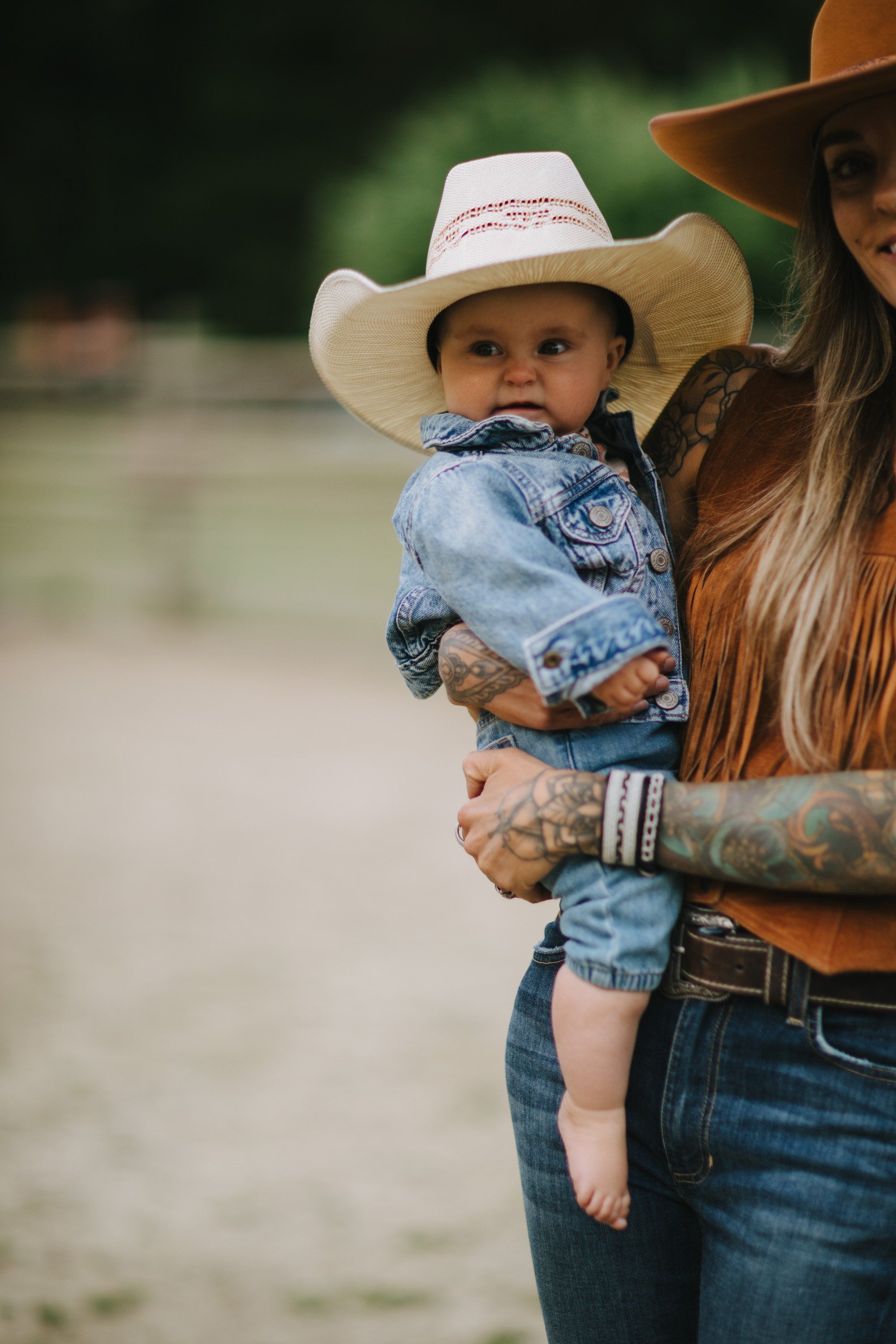 Woman in cowboy hat holding toddler in denim jacket and white hat on rural road, golden hour light
