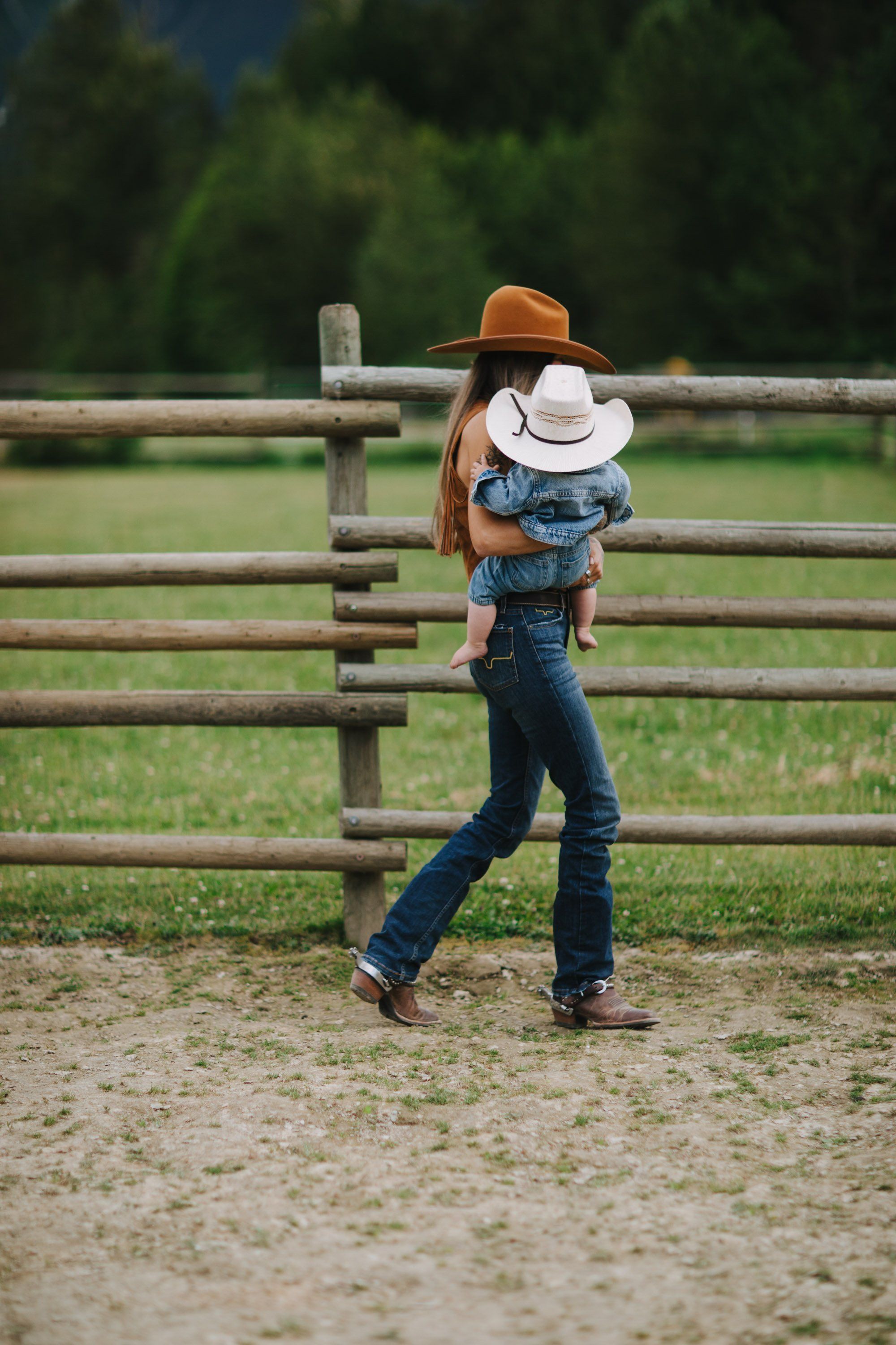 Woman in brown hat carrying child in white hat stands by wooden ranch fence on sunny day