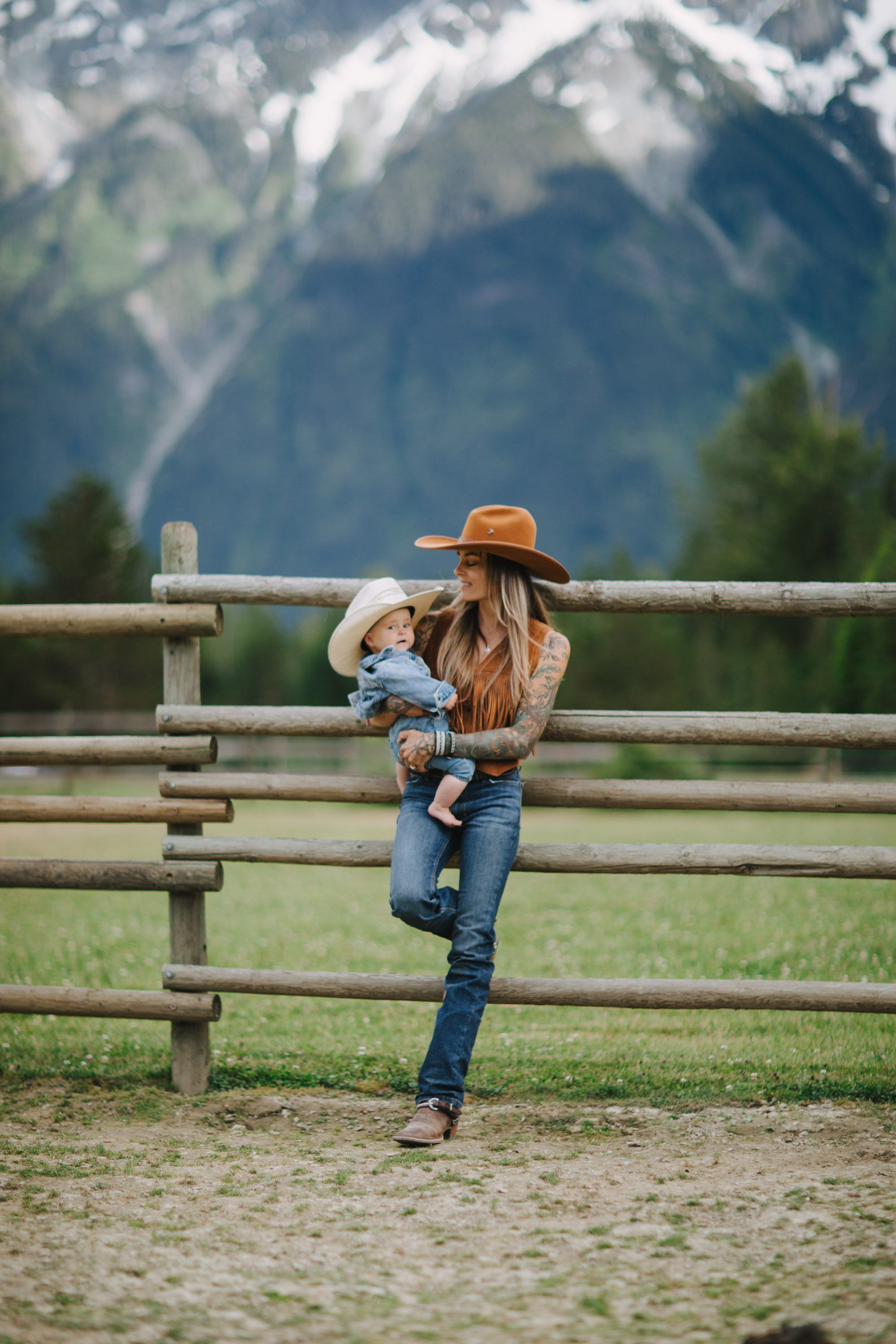 Mother holding toddler on wooden ranch fence with snow-capped mountains in soft focus behind them