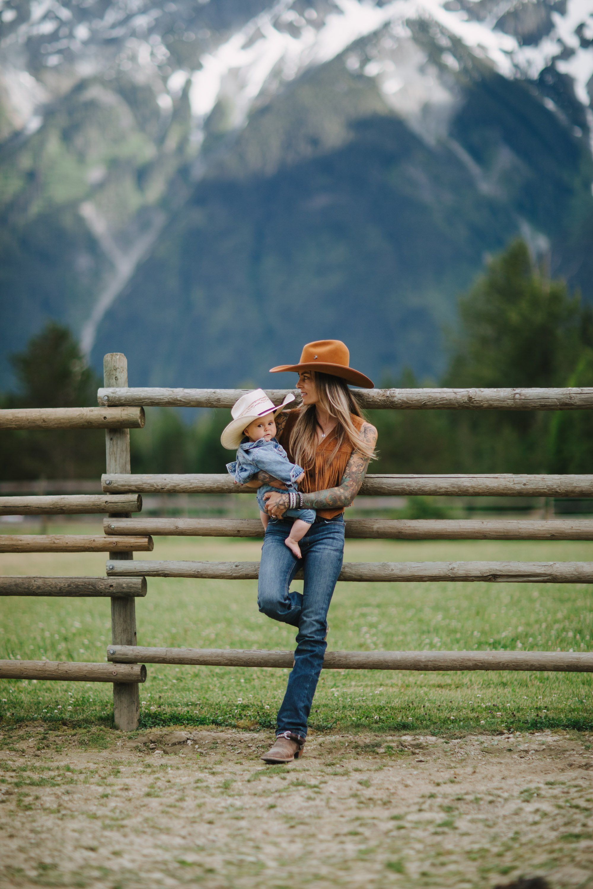 Woman in cowboy hat holding toddler on wooden ranch fence with snow-capped mountains in soft focus behind