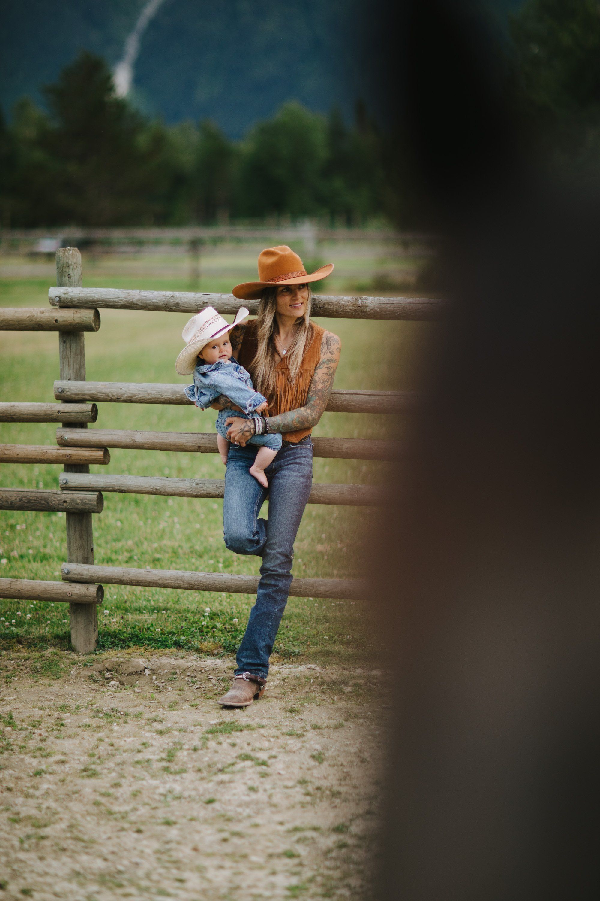 Woman in orange hat holding child by wooden ranch fence under bright afternoon light