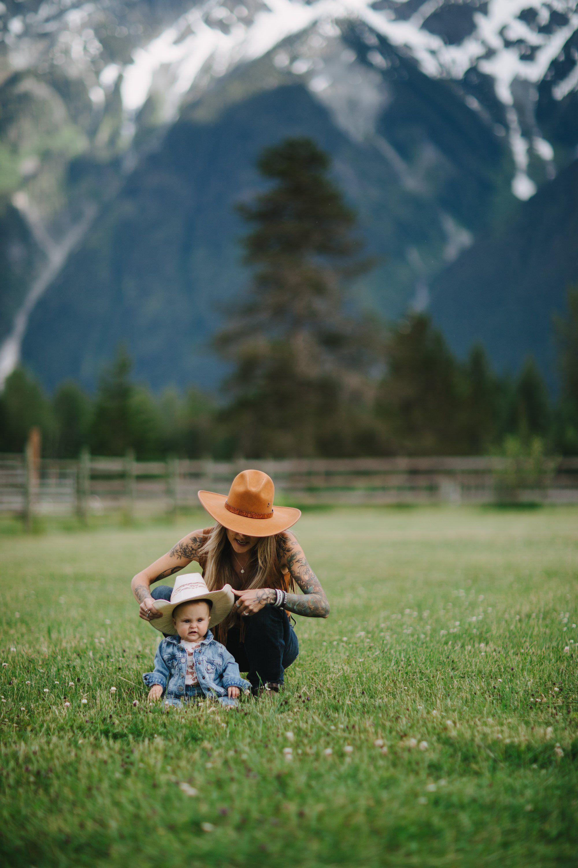 Adult and toddler in cowboy hat sit together in sunlit meadow with snow-capped mountains behind