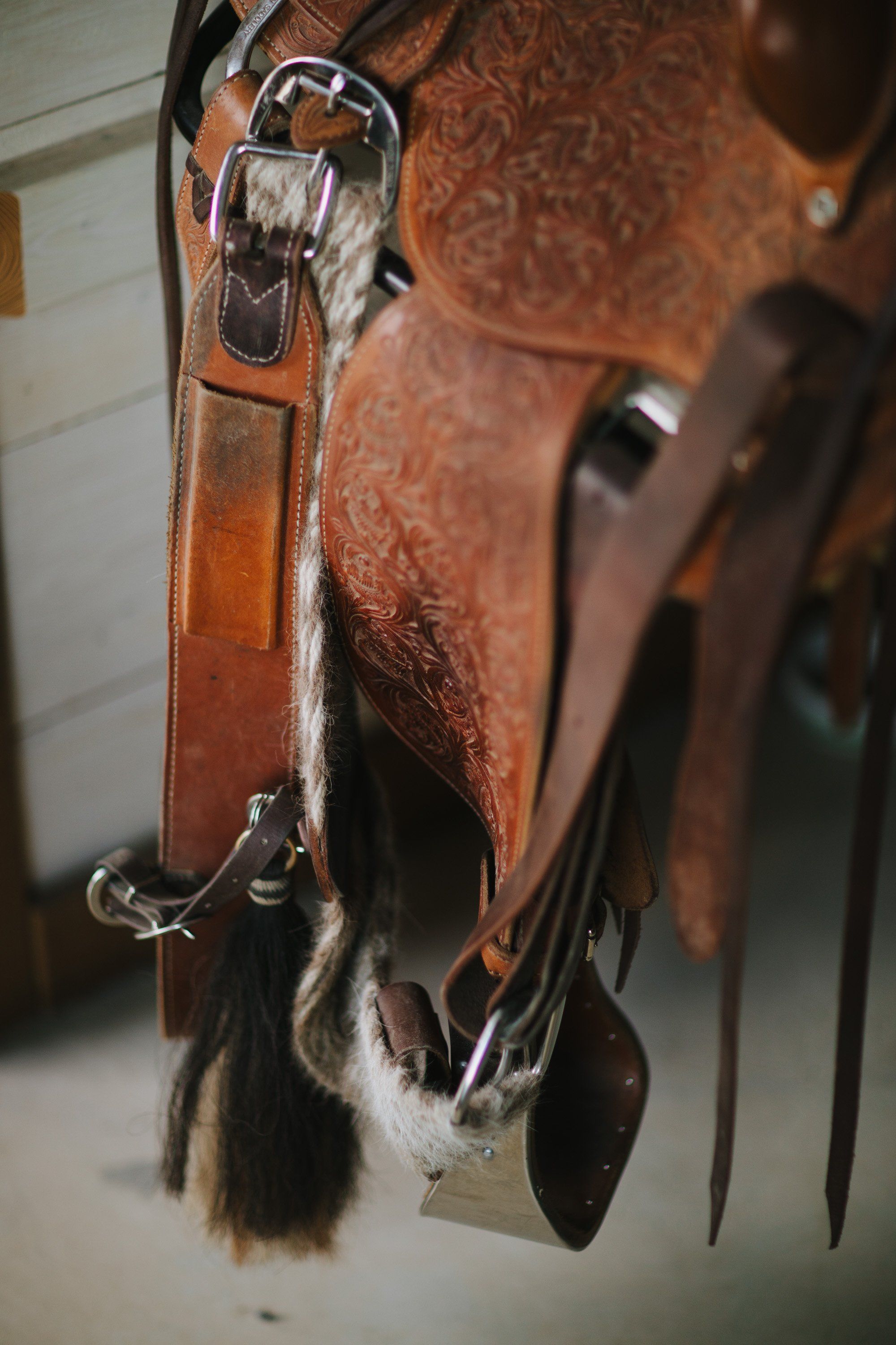 Worn leather saddle with tooled details and hair-covered stirrups hanging against a neutral wall