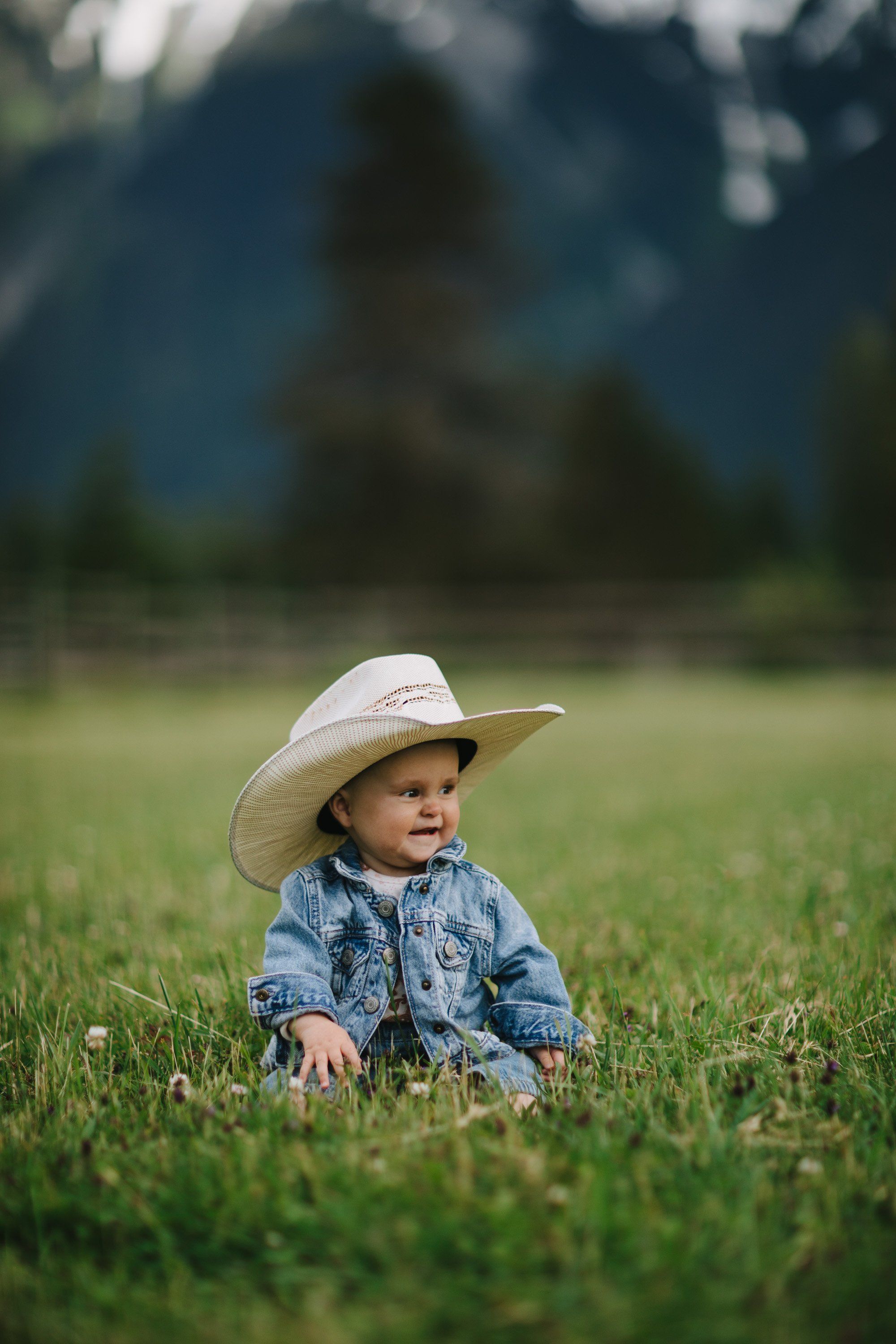 Toddler in denim shirt and cream cowboy hat sits in green meadow with mountain peaks and forest backdrop