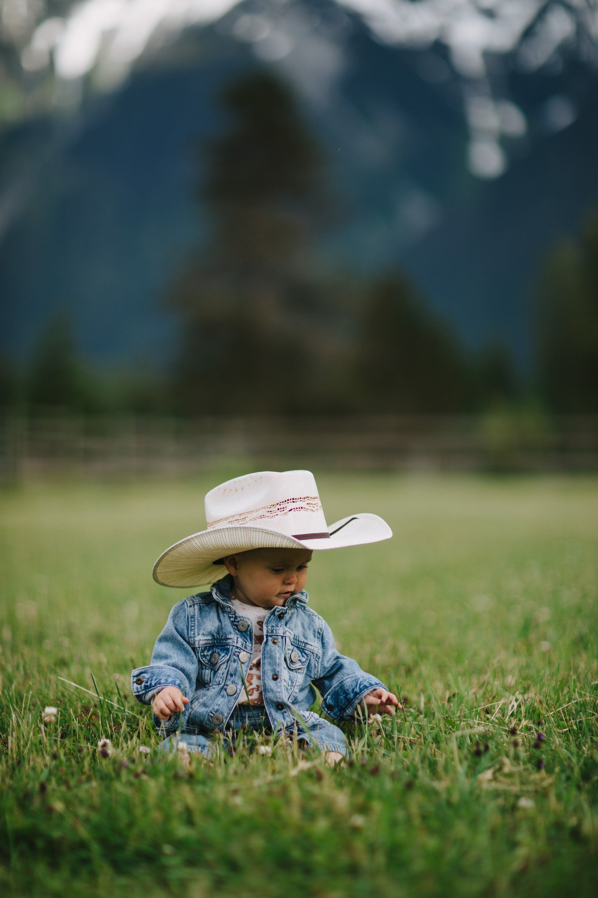 Toddler in cowboy hat and denim jacket sitting in a grassy meadow with snow-capped mountains in the soft-focus background