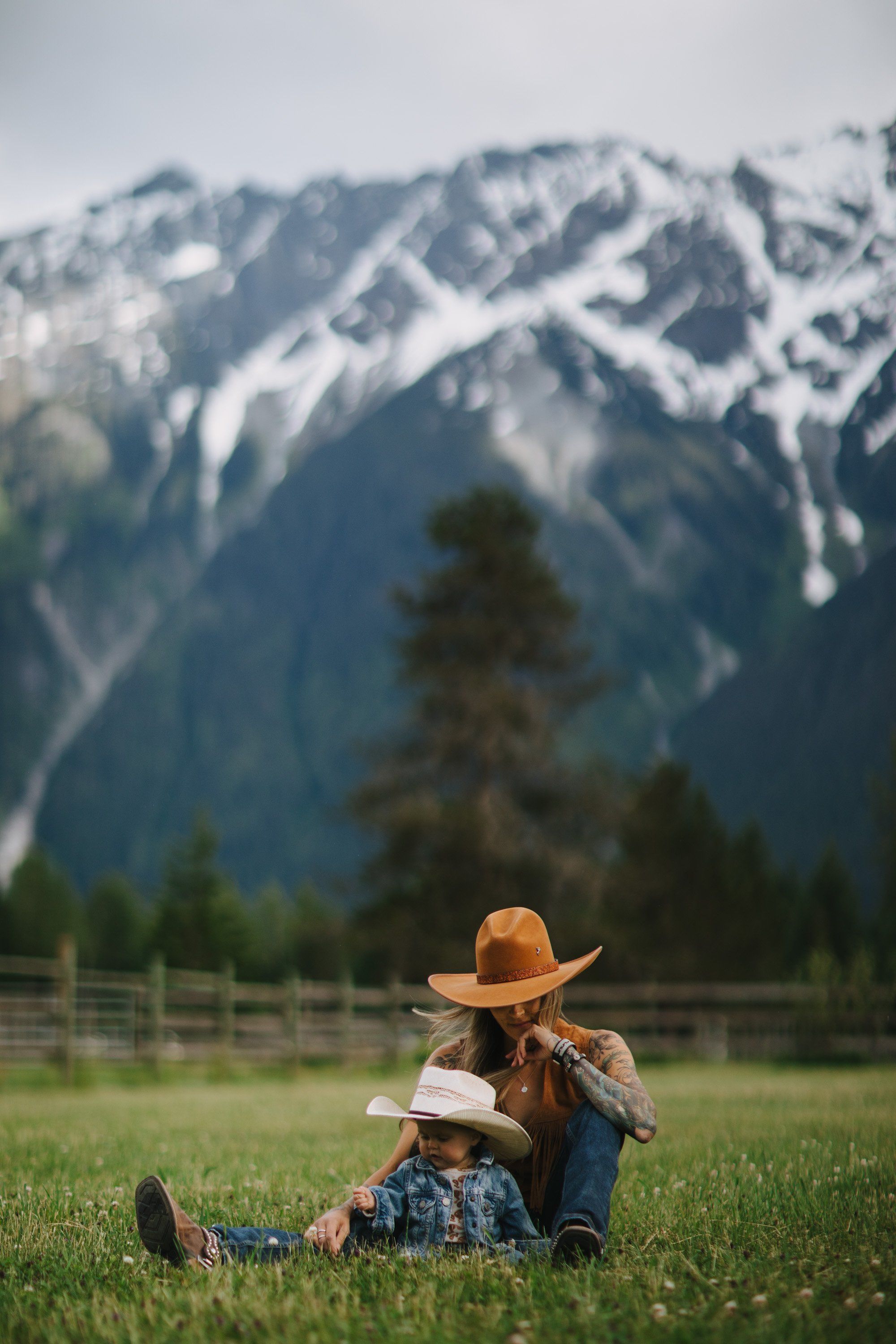 Adult and child sit together in meadow grass wearing cowboy hats, snow-capped mountains rising behind them