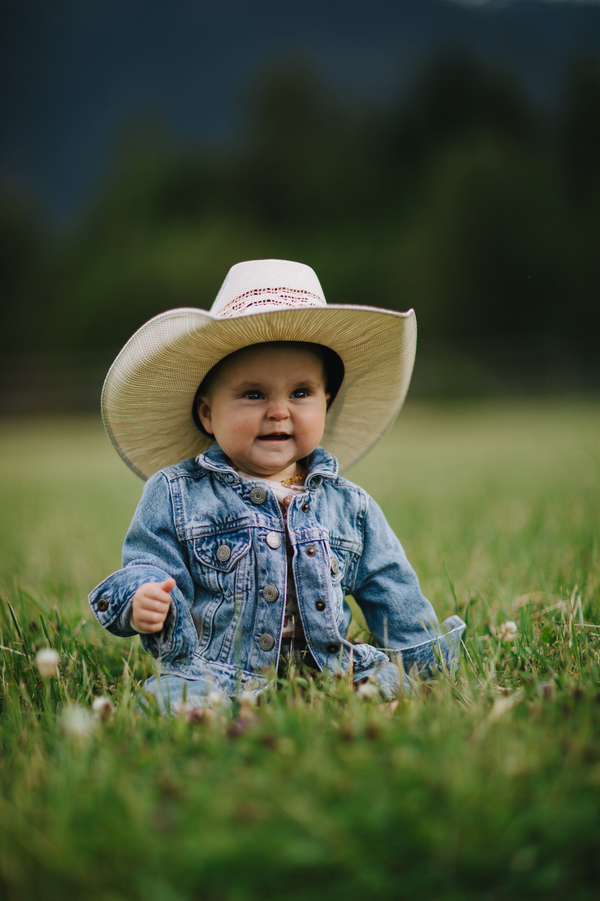 Toddler in denim shirt and oversized straw cowboy hat sits in grass with blurred green trees behind