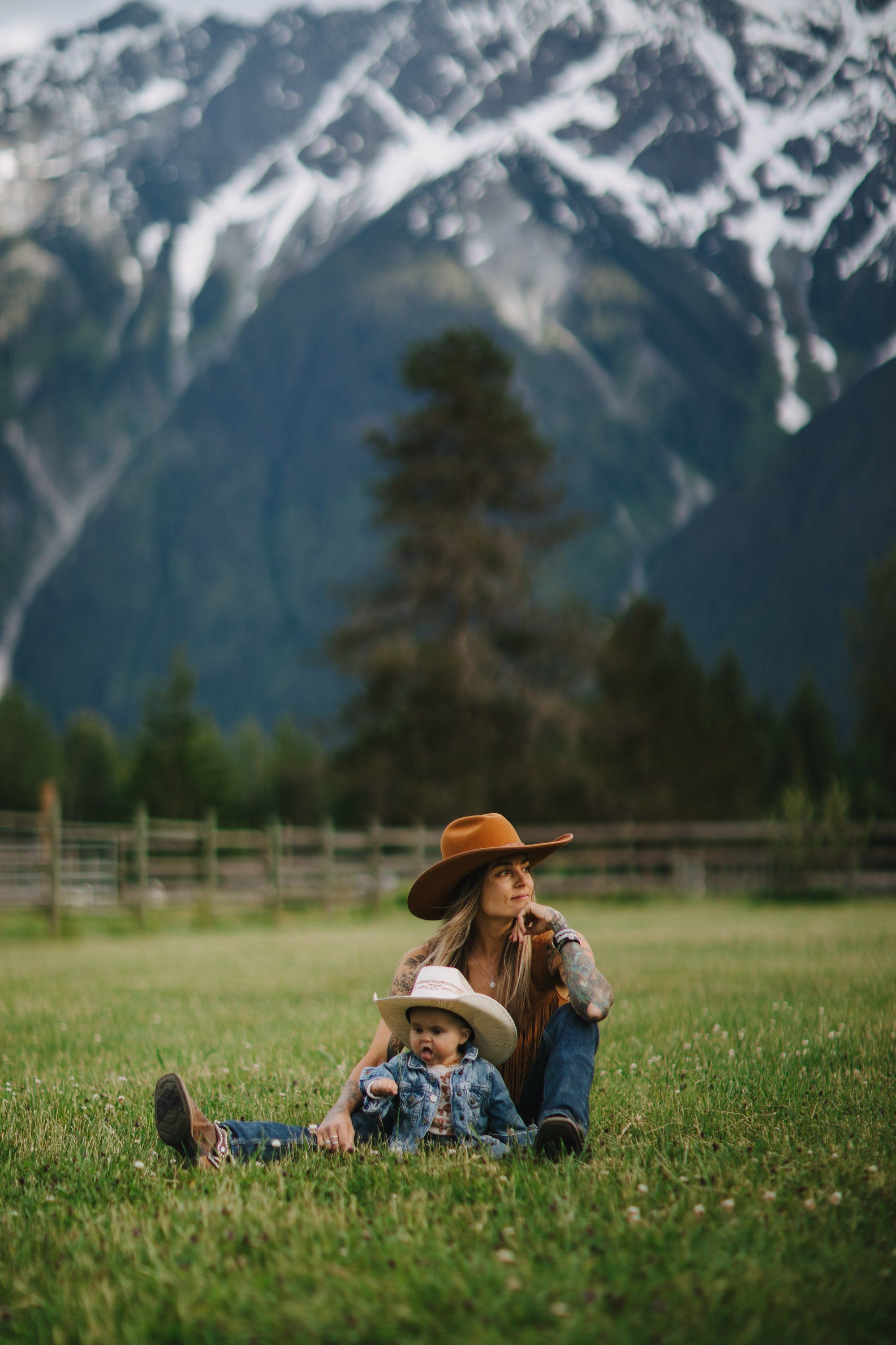 Mother and child sit together on a sunlit meadow with snow-capped mountains and evergreen trees behind them