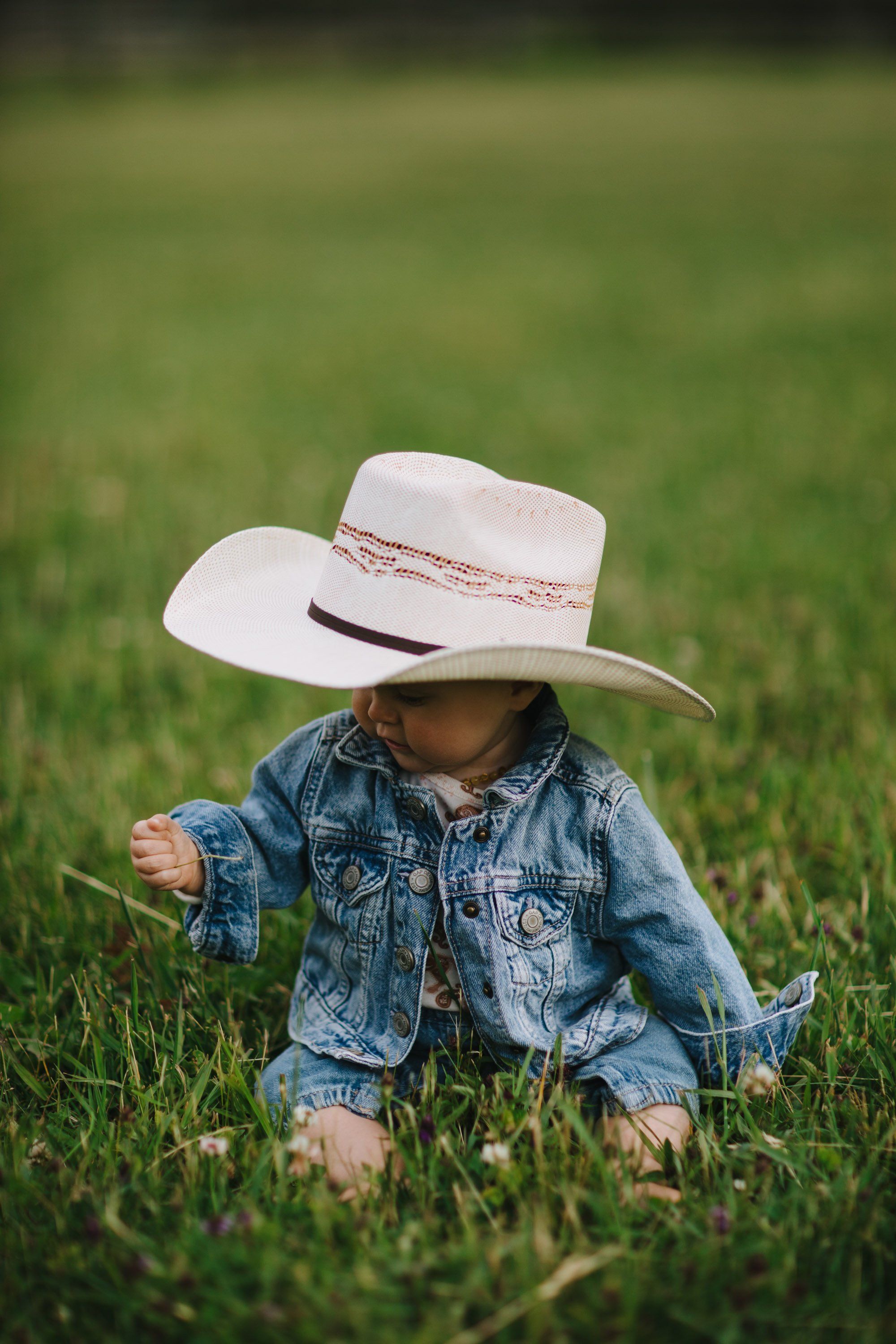 Toddler in denim shirt and cream cowboy hat sits on grass, soft afternoon light filtering through blurred green background