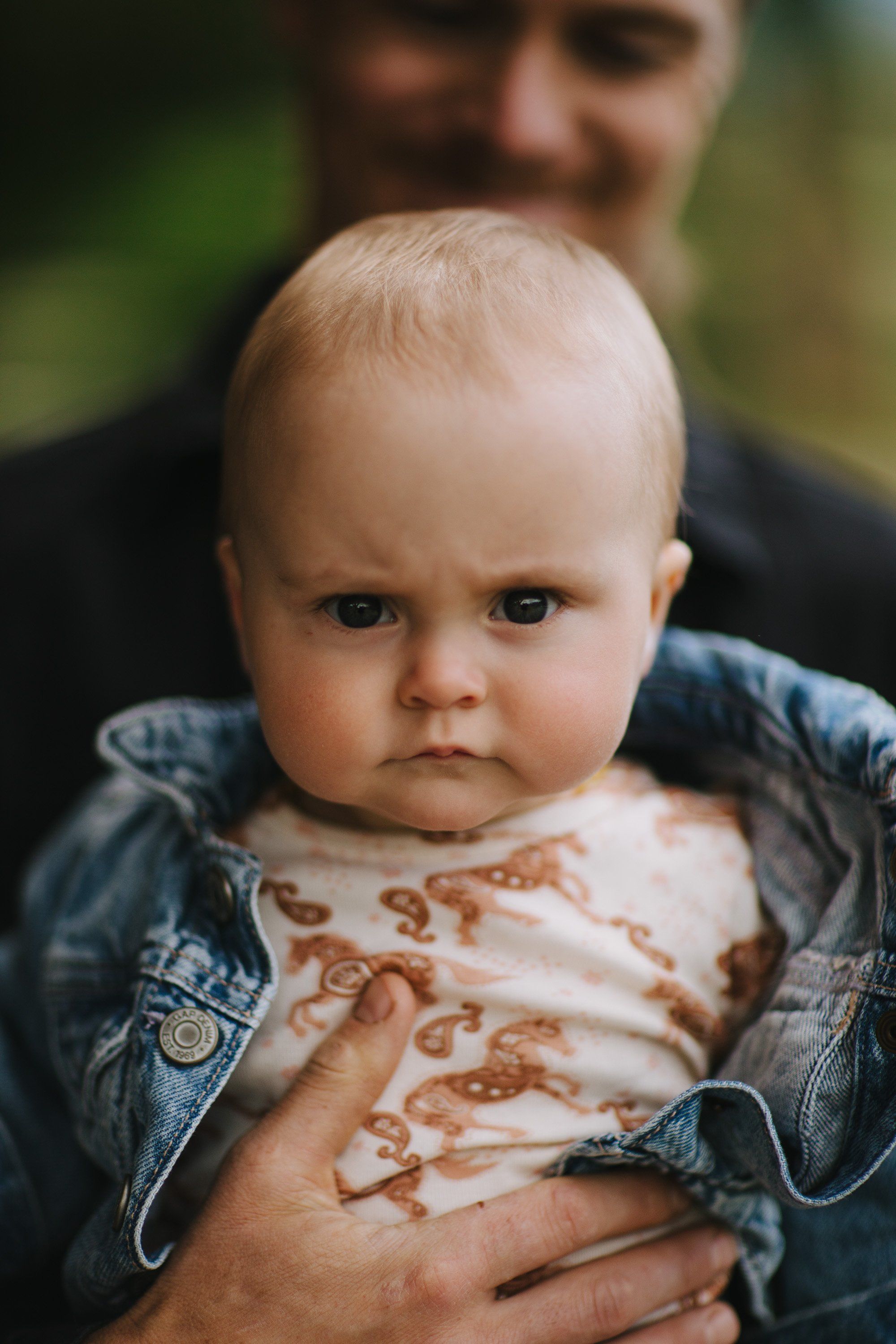 Blond toddler with serious expression held by adult in denim jacket, soft natural light, green background