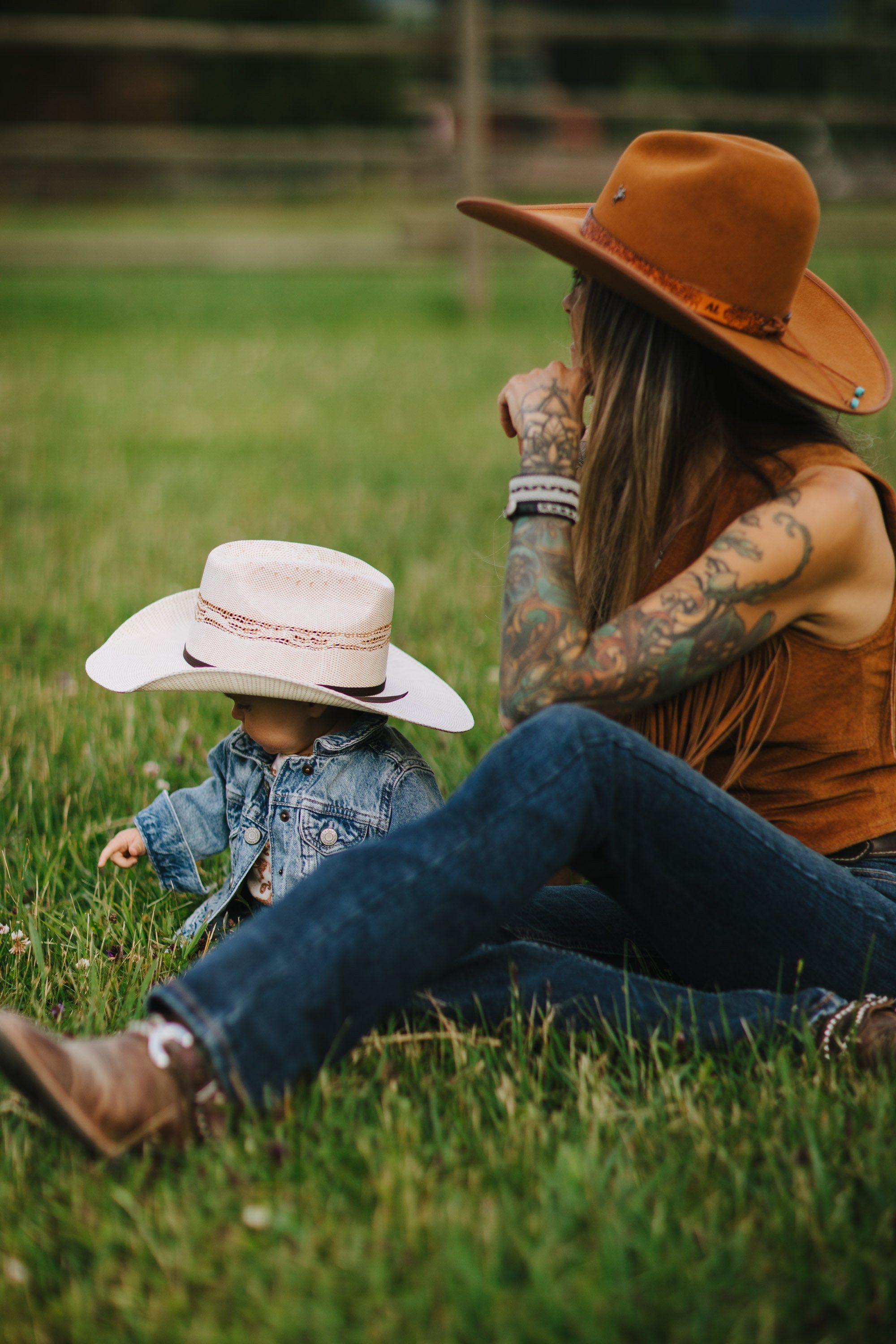 Woman and young child wearing matching cowboy hats sit together on grass, bathed in golden afternoon light