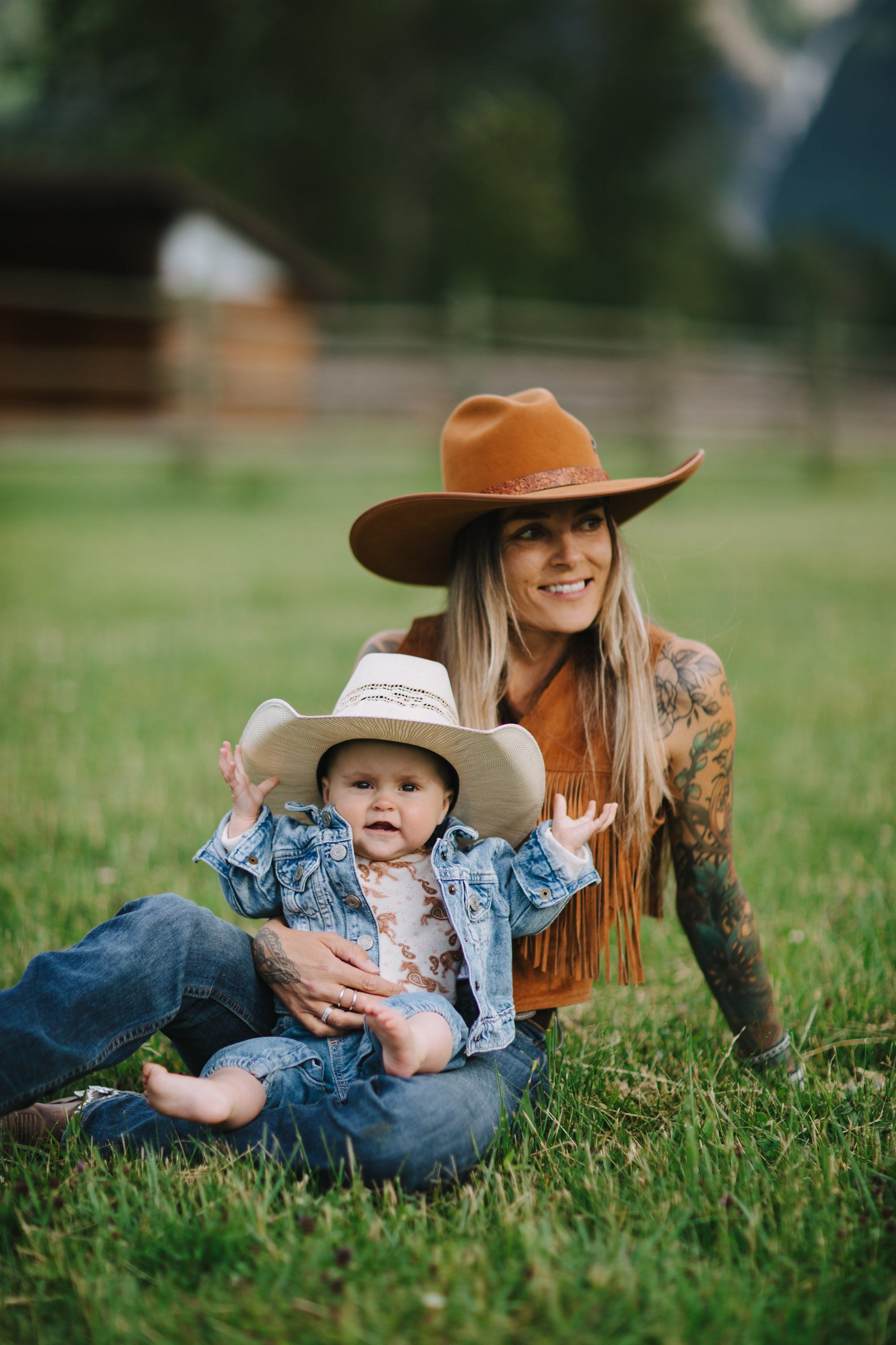 Woman and toddler in cowboy hats sit on grass, she in orange felt and fringe, child in denim, golden hour light filtering through trees behind them