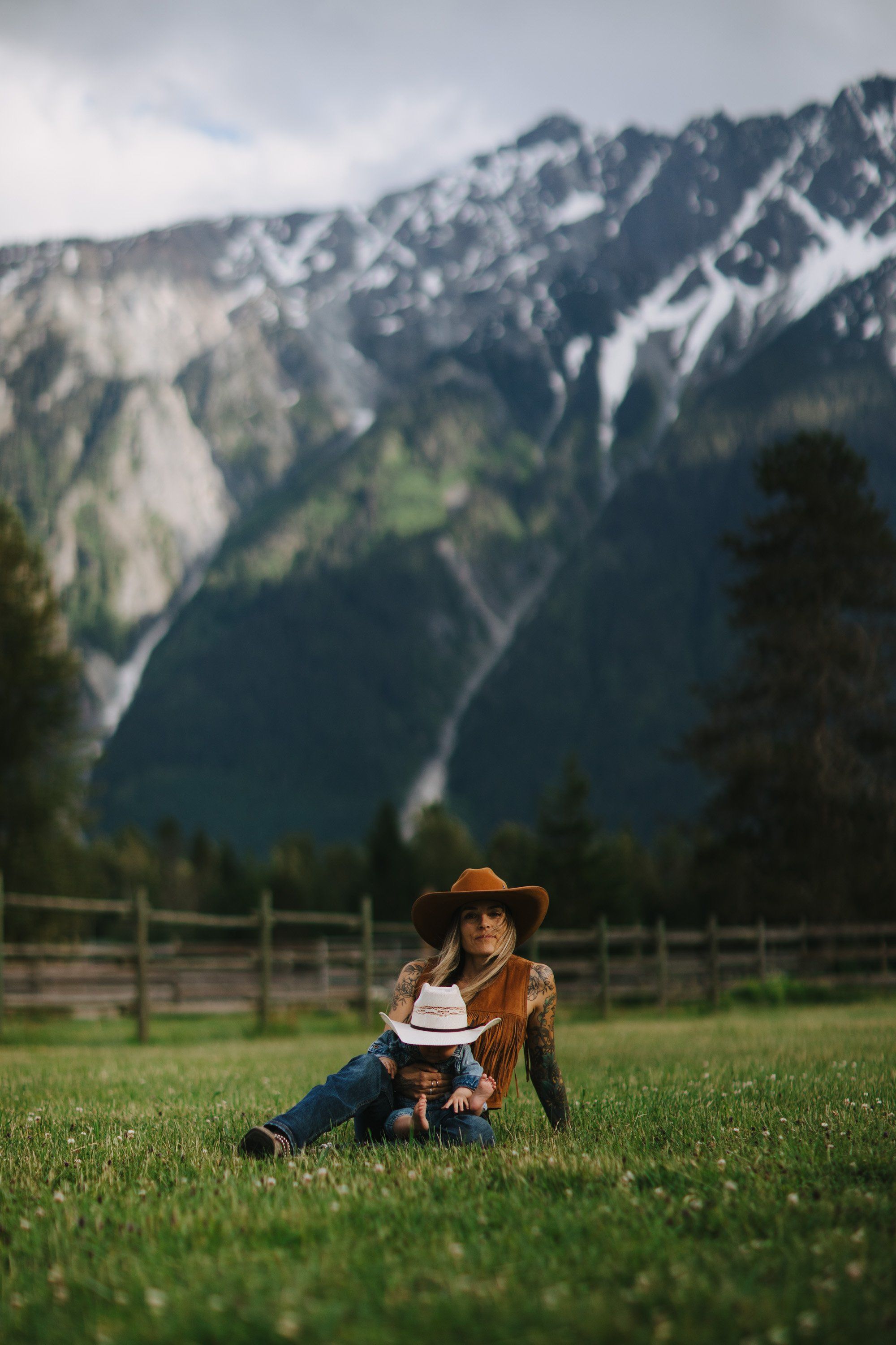 Child in cowboy hat sits in green meadow with snow-capped mountains and wooden fence behind