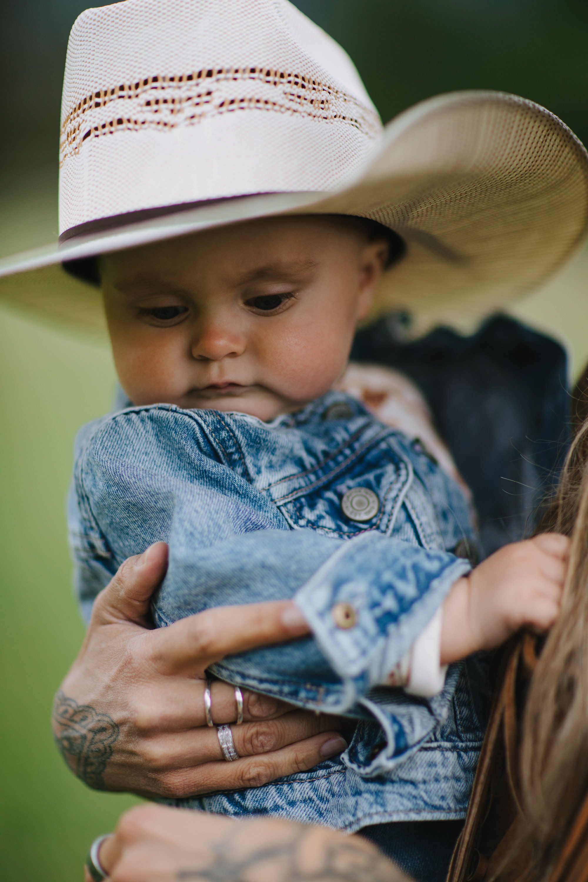 Toddler in denim shirt and cream cowboy hat held by adult against soft green background