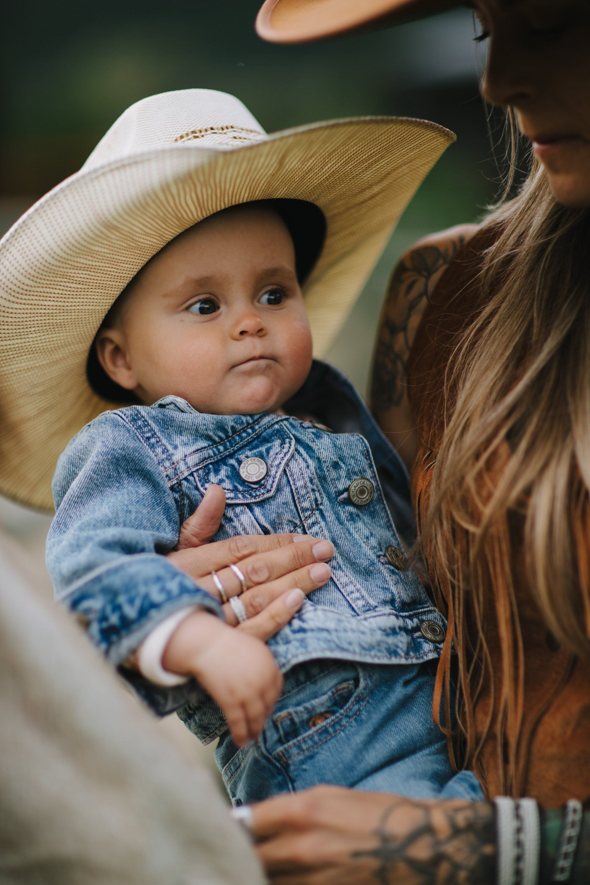 Toddler in denim shirt and cream cowboy hat held by parent with long brown hair and visible tattoo, soft natural light, intimate family moment