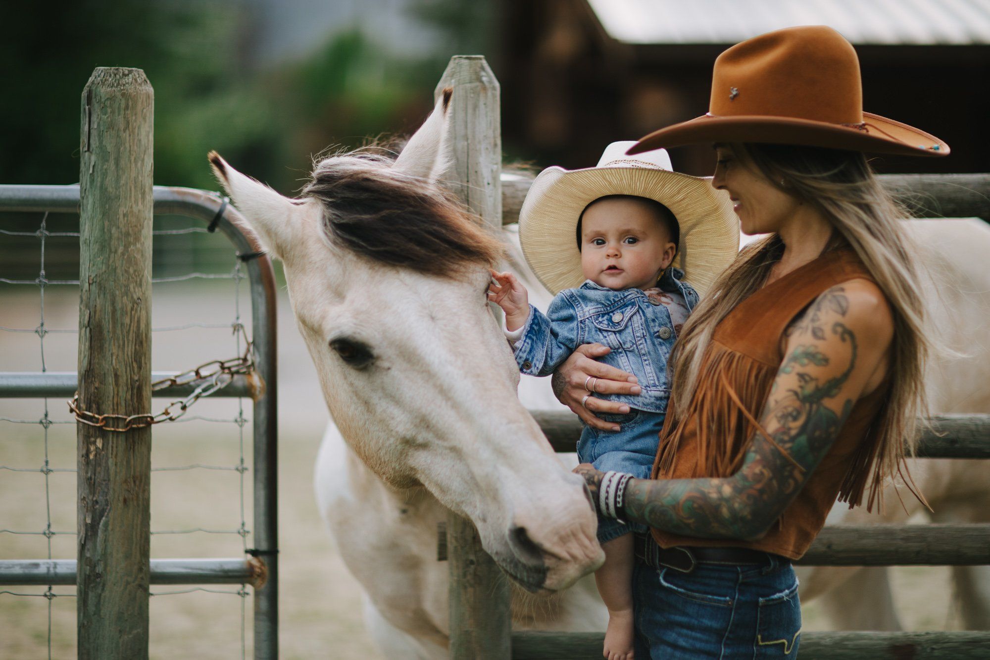 Woman in brown cowboy hat and fringe jacket holds baby in straw hat while feeding white horse at wooden ranch fence