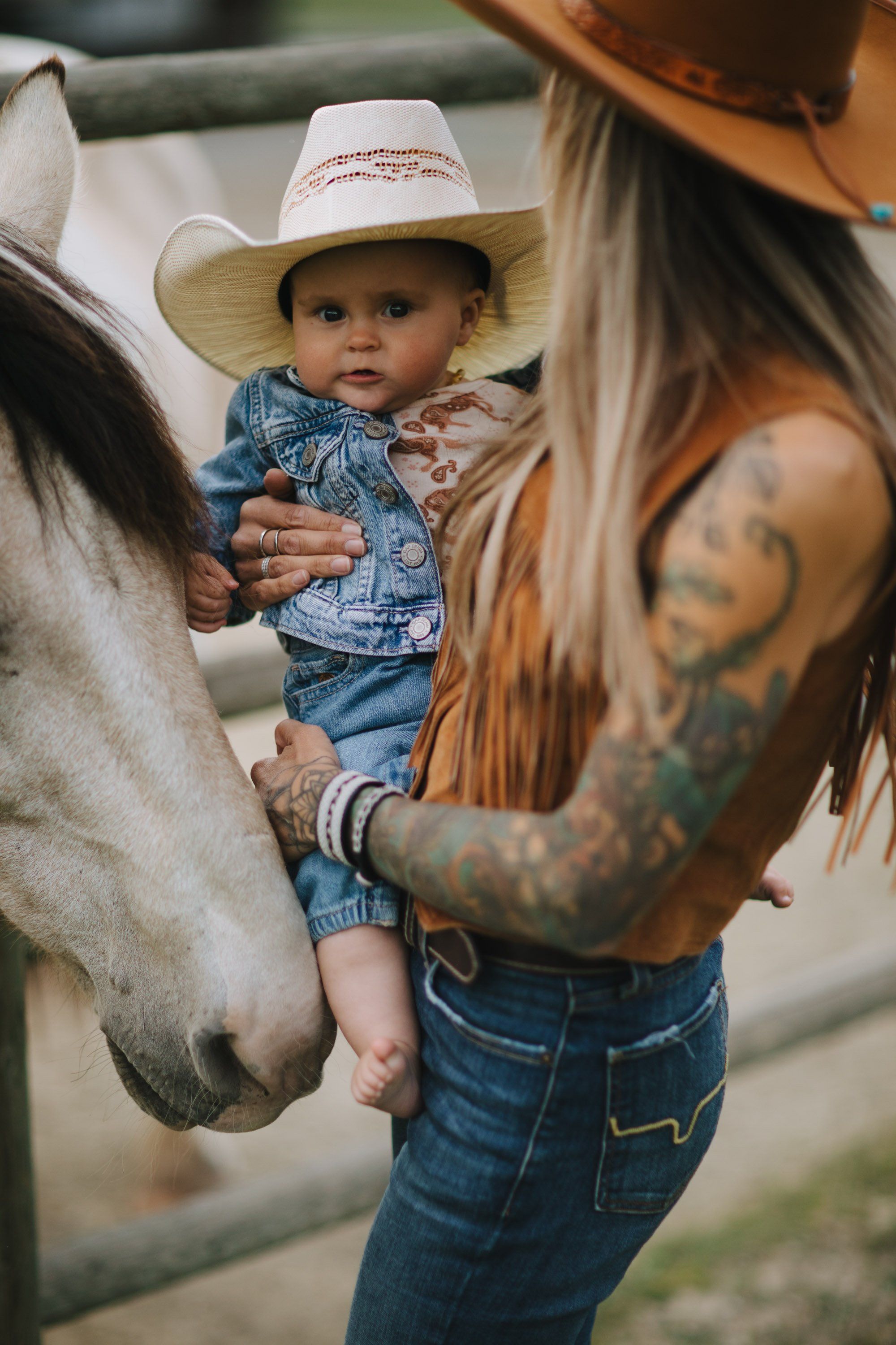 Woman with visible arm tattoos holding a toddler in cowboy attire next to a horse in golden afternoon light