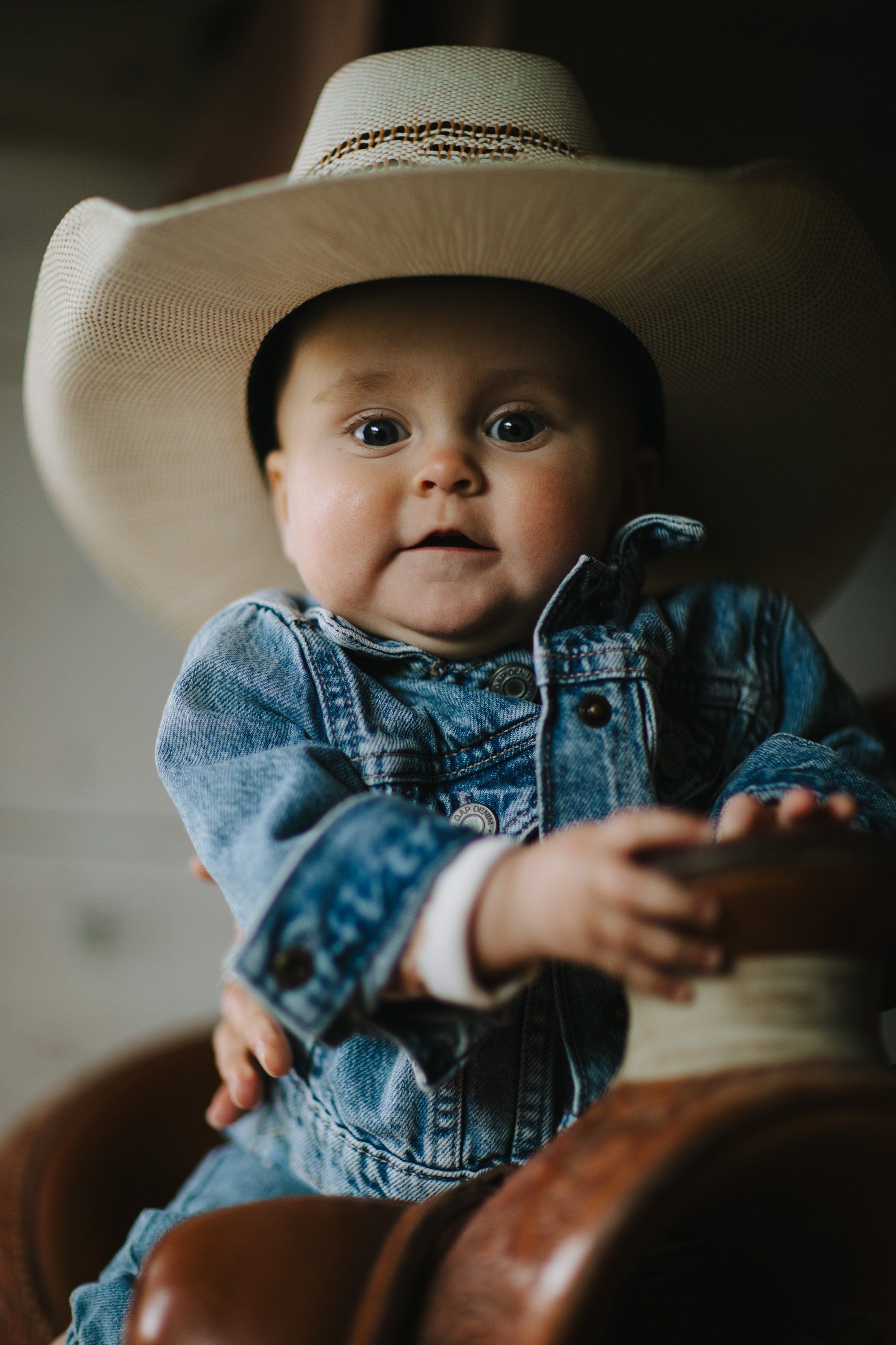 Baby in denim shirt and cream cowboy hat sits on brown leather, reaching toward camera with soft natural light
