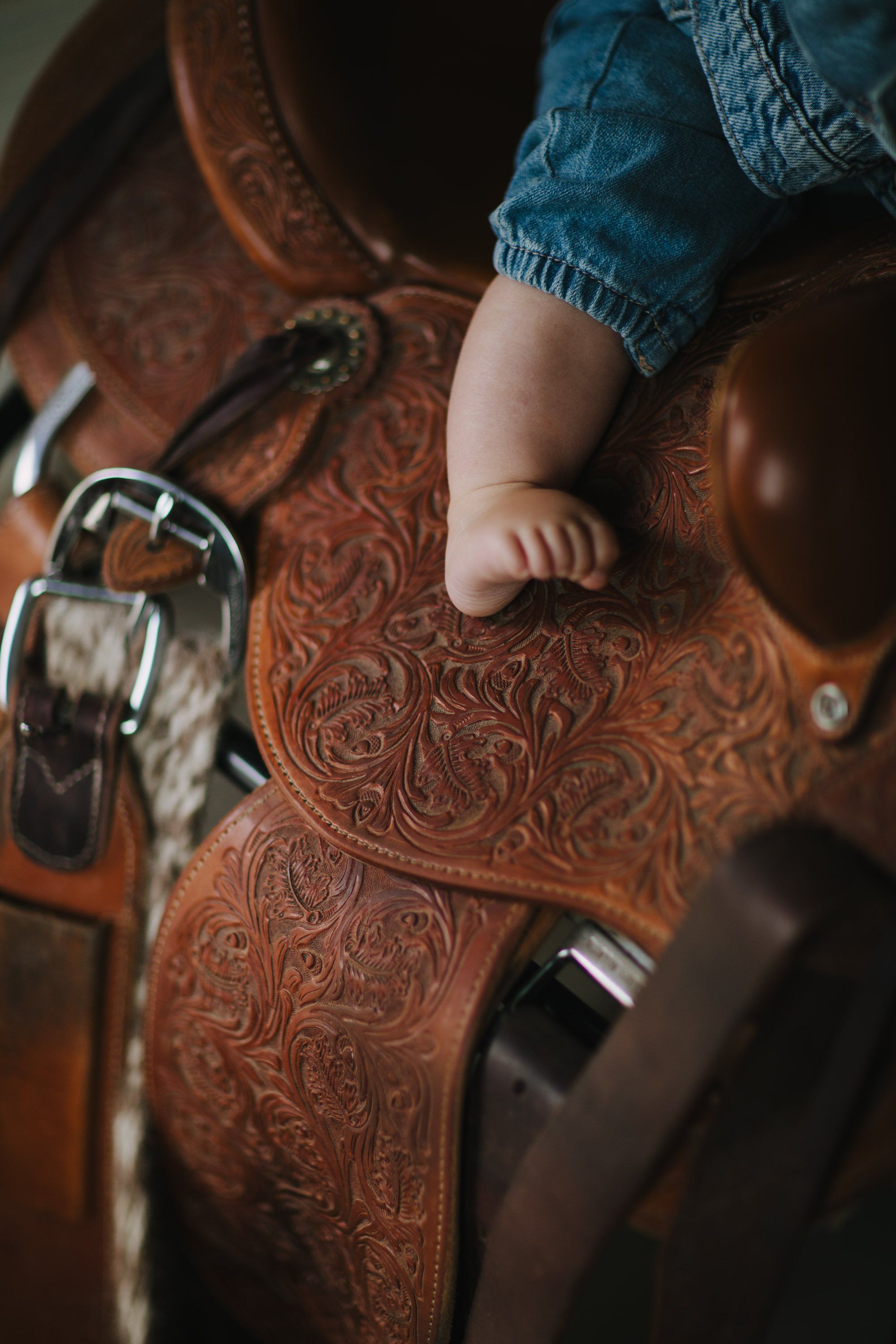 Baby's bare foot rests on ornate brown leather saddle with decorative tooling and metal fittings