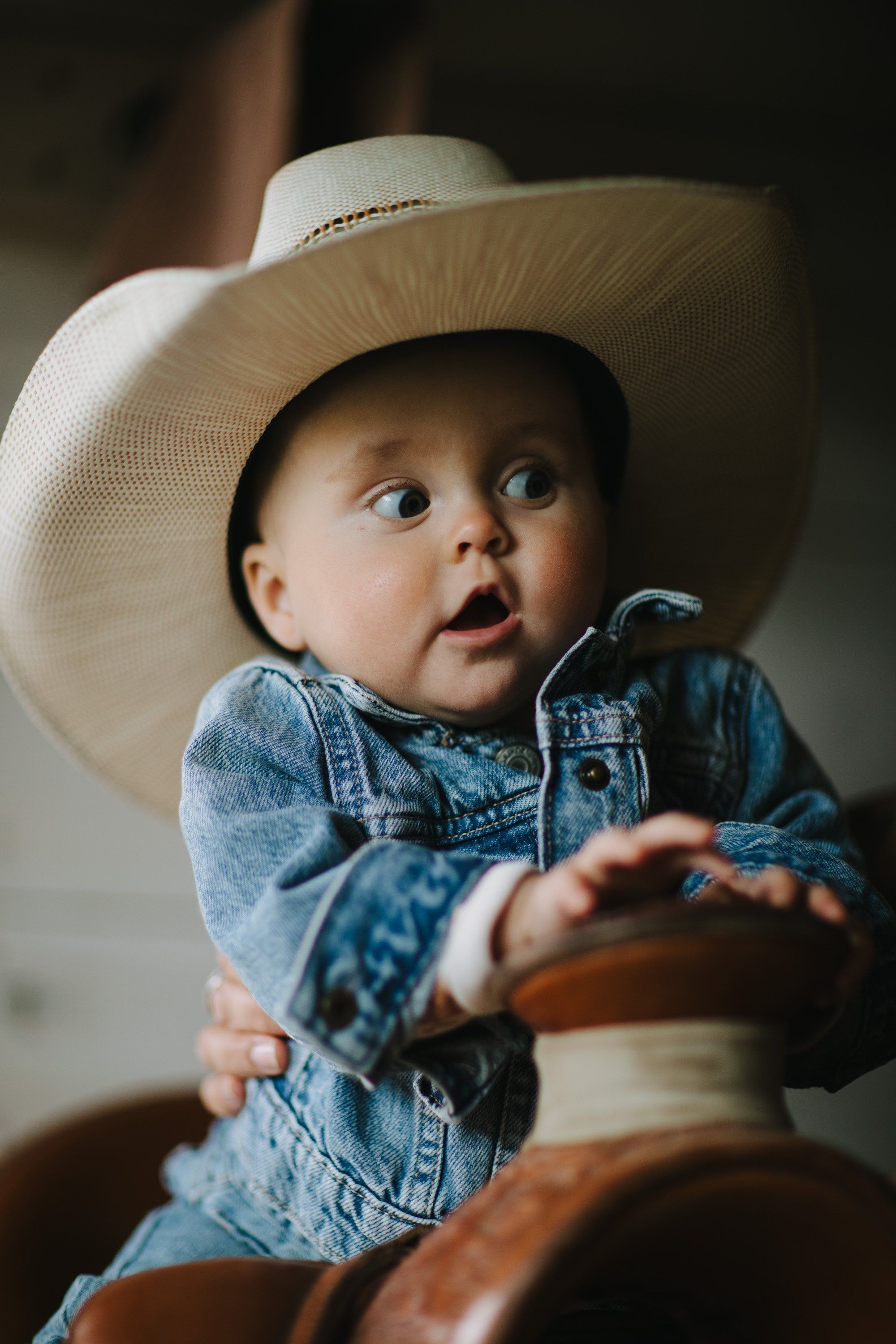 Surprised baby in oversized cream cowboy hat and denim shirt sits on brown leather saddle, mouth open in wonder