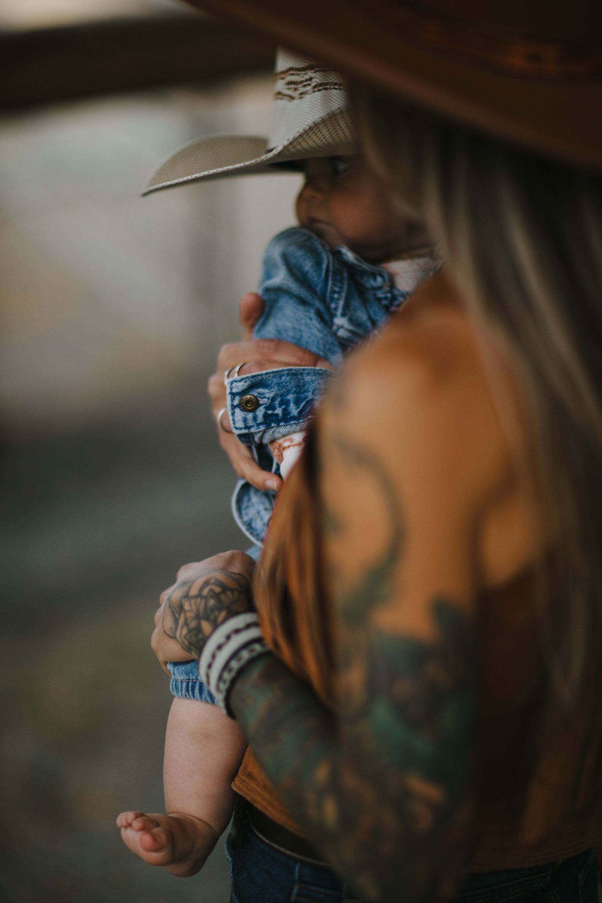 Shirtless father wearing cowboy hat holds young child in denim shirt against warm, blurred background in golden afternoon light
