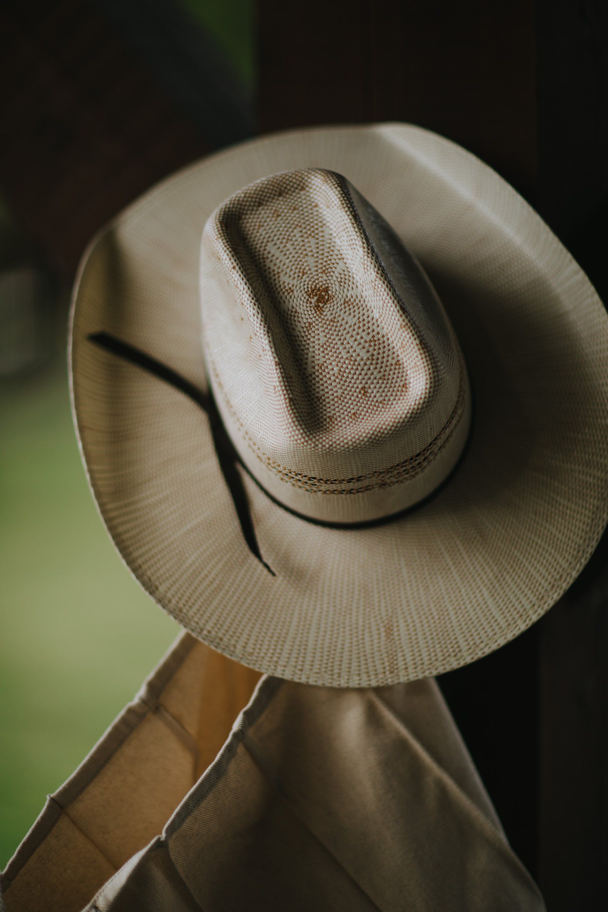 Cream cowboy hat tilted back on a person's shoulders against soft green and dark backgrounds, intimate detail of western wear