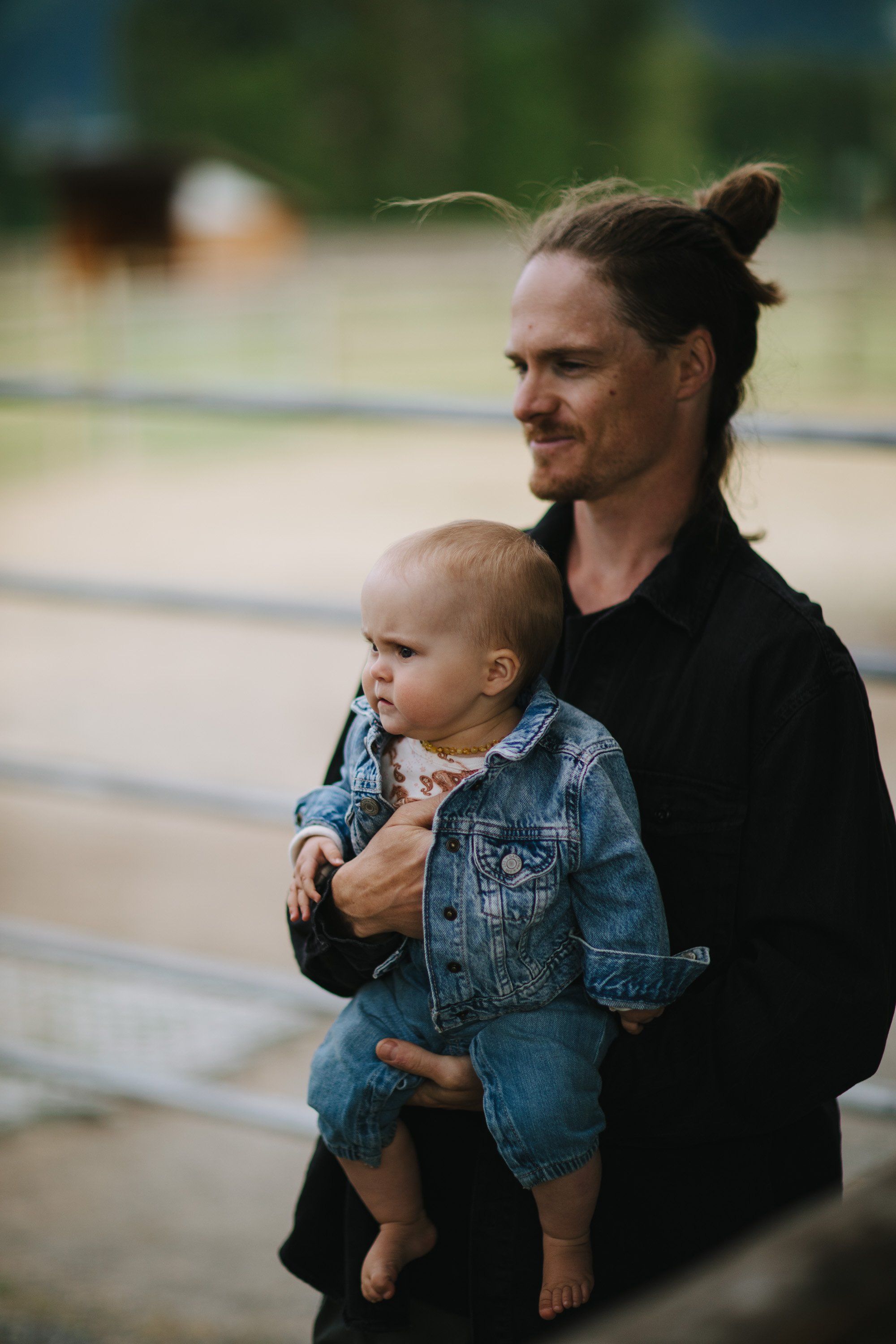 Father with man bun holding toddler in denim jacket, soft natural light, blurred background with trees