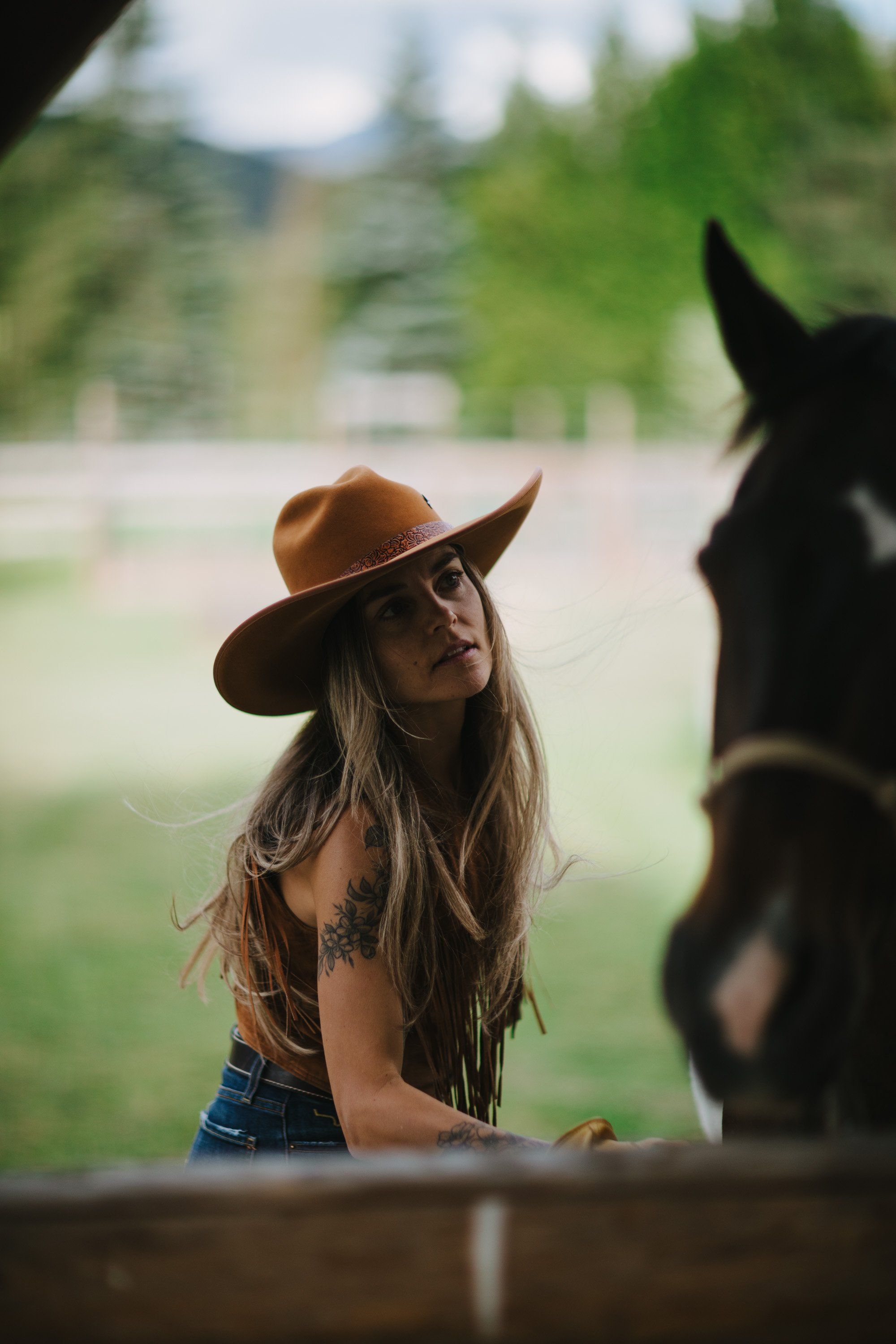 Woman in cowboy hat and fringe jacket gazes at dark horse beside wooden fence in pastoral mountain landscape