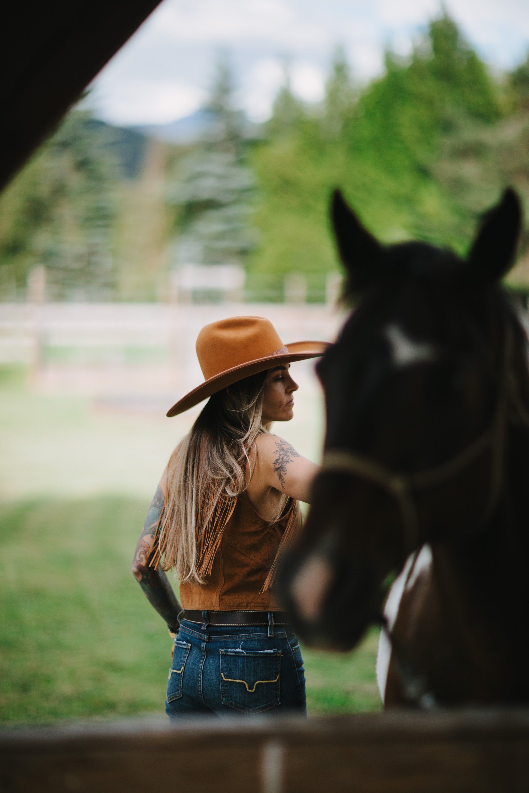 Woman in brown cowboy hat and tan jacket stands beside dark horse in wooden stable, with forested mountains visible through the opening behind her