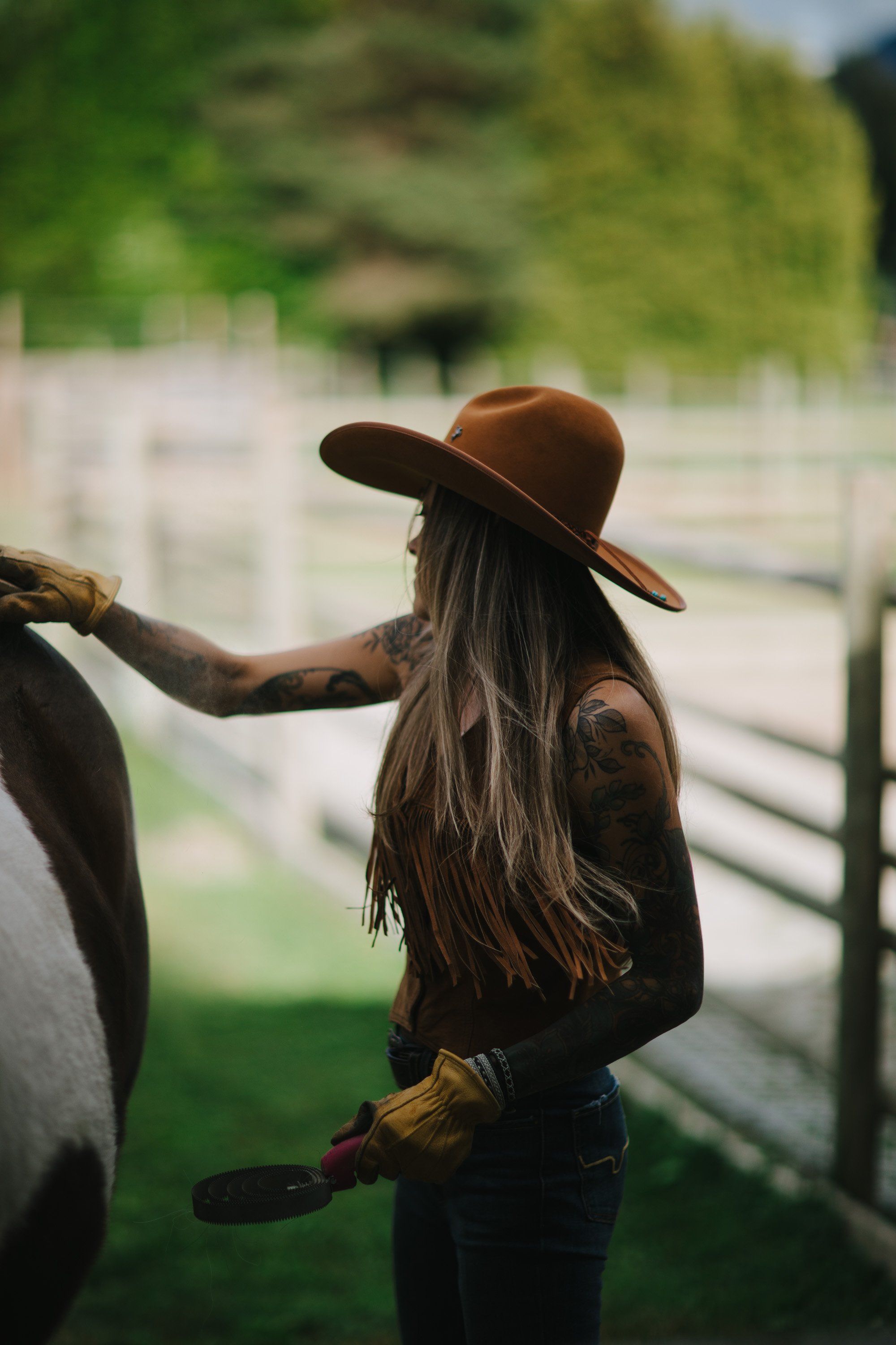 Woman in brown cowboy hat and dark jacket feeding a horse, soft afternoon light filtering through trees in rural setting
