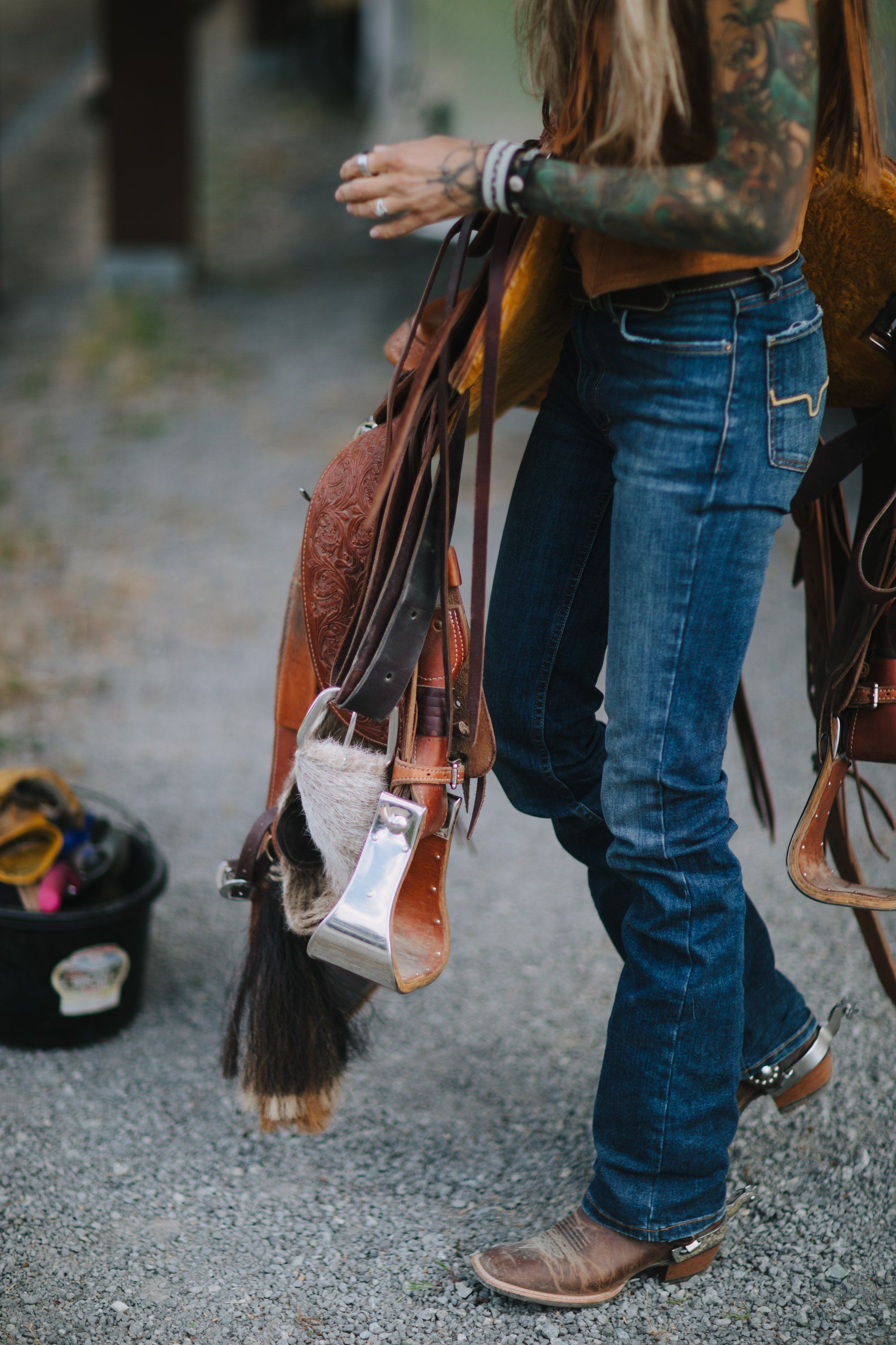 Woman with tattoos holding a cognac leather bag against her denim-clad leg in soft outdoor light