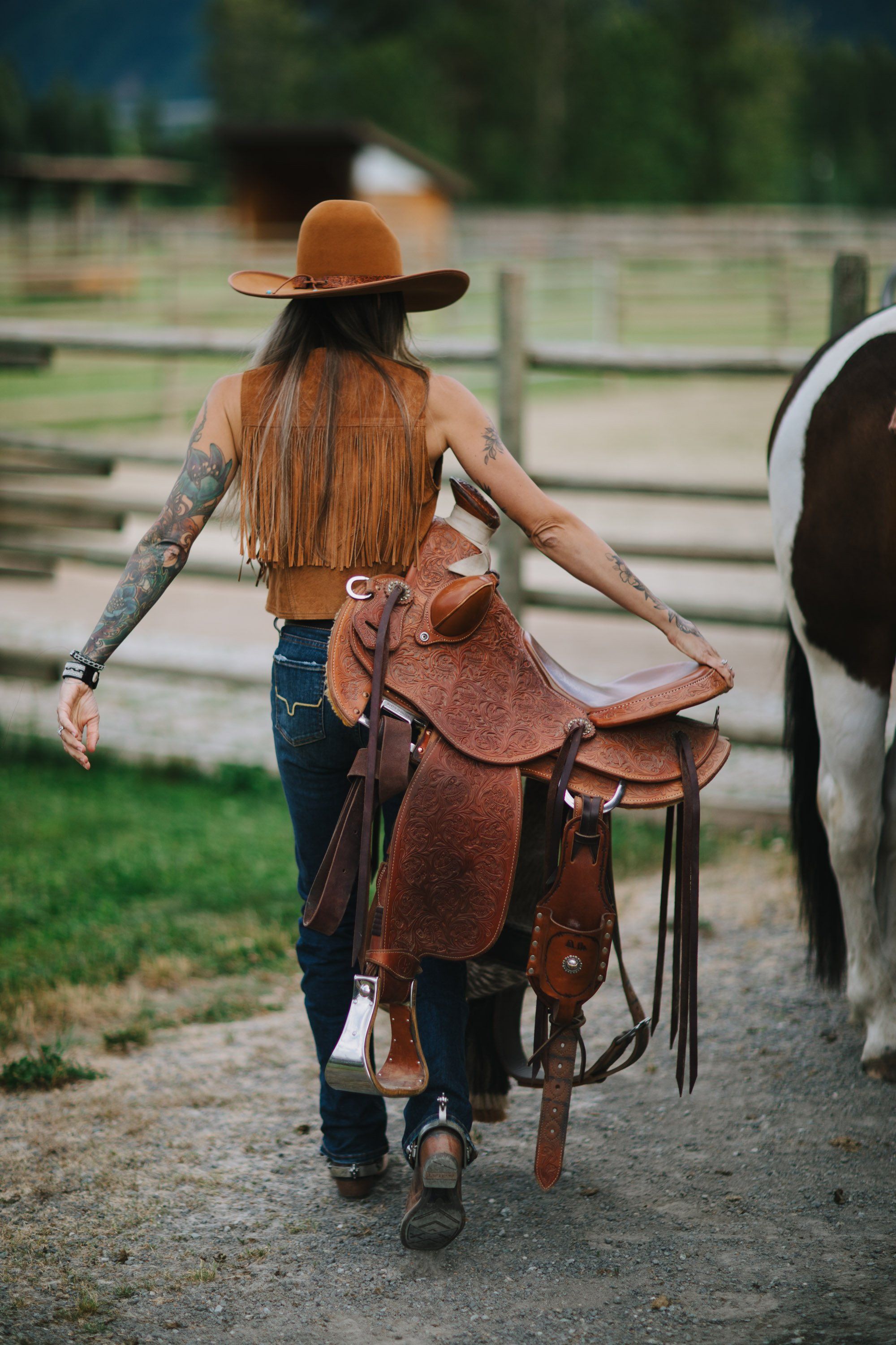 Woman with long auburn hair and tattoos carries a leather saddle toward a horse in a sunlit ranch setting