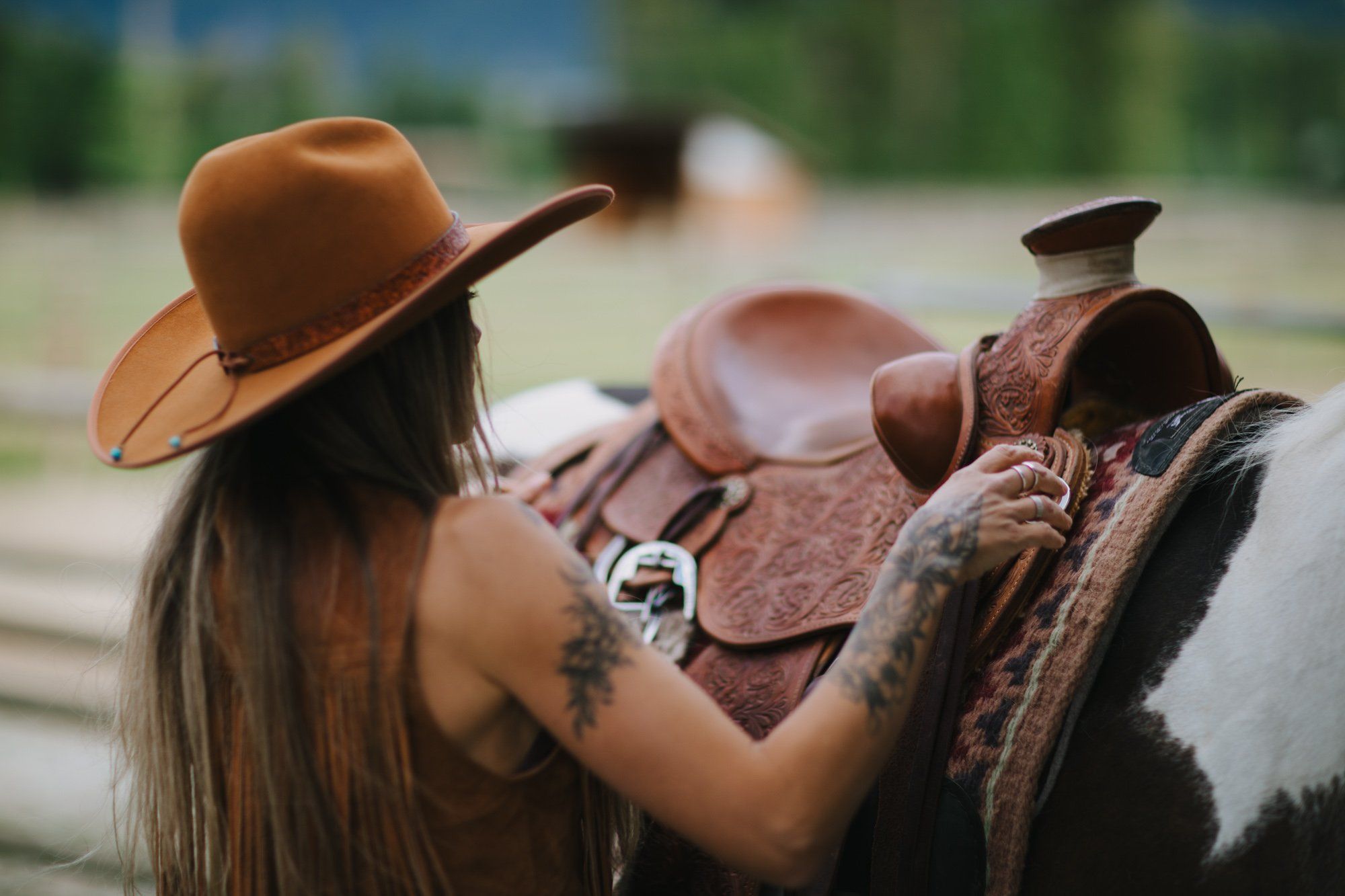 Woman in rust cowboy hat adjusting ornate western saddle on horse, intimate moment in pastoral setting