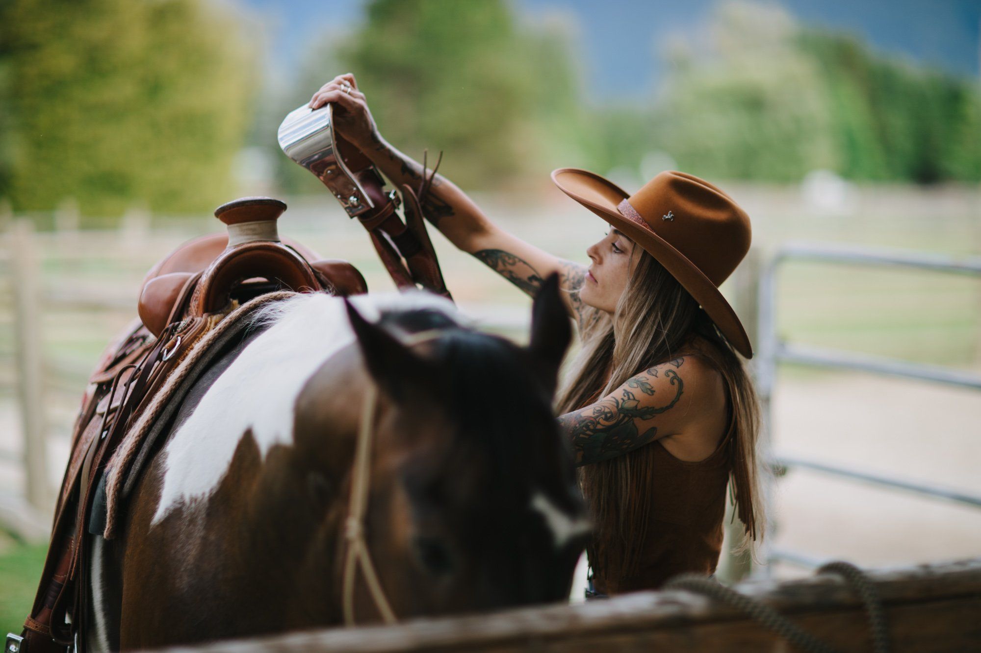 Cowboy adjusting saddle on dark horse with long hair, wide-brimmed hat, pastoral ranch setting in soft afternoon light